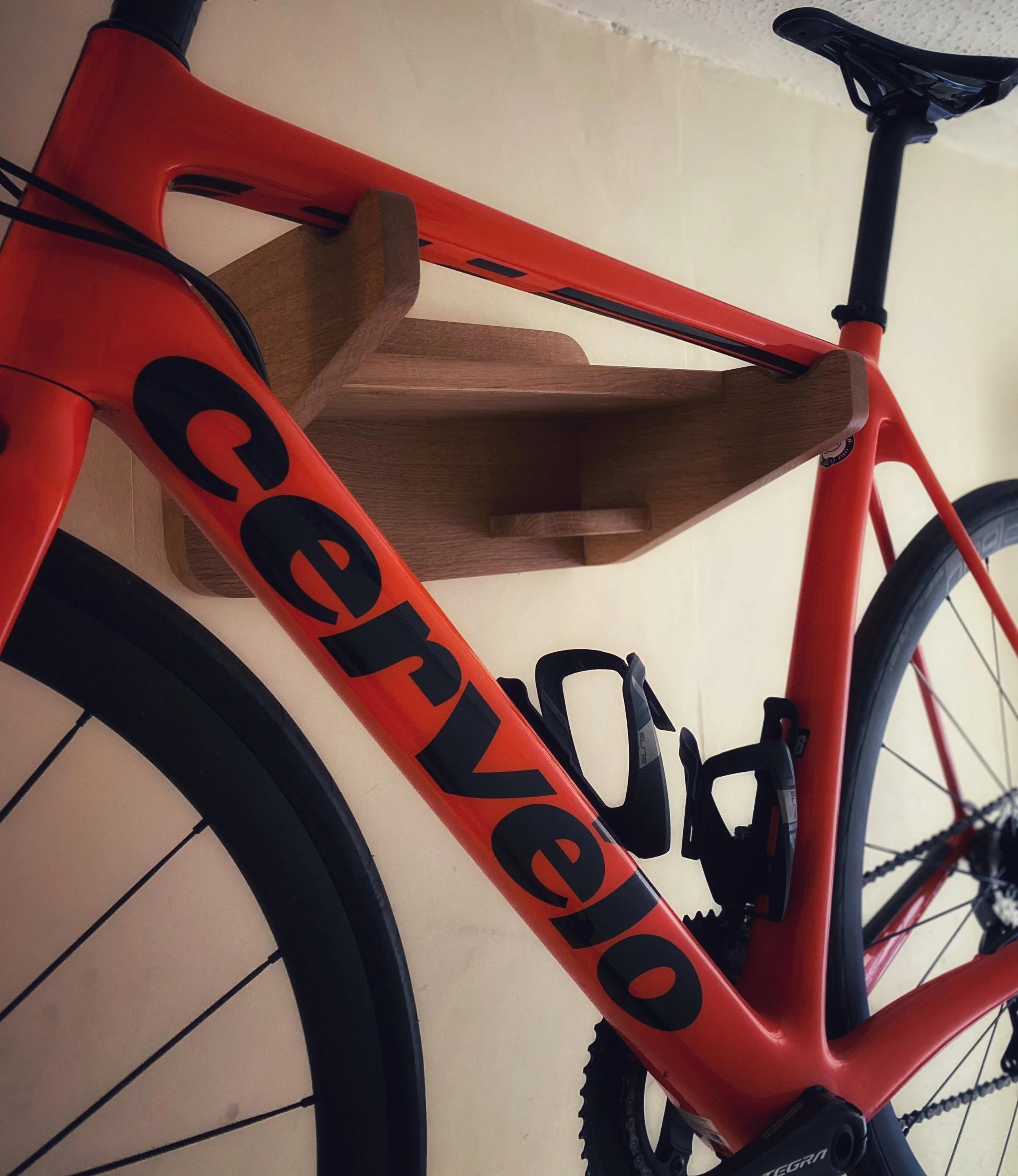 A red bicycle with black accents, placed on a light-colored tile floor, with a wooden stand supporting the frame, and part of a wall visible in the background.