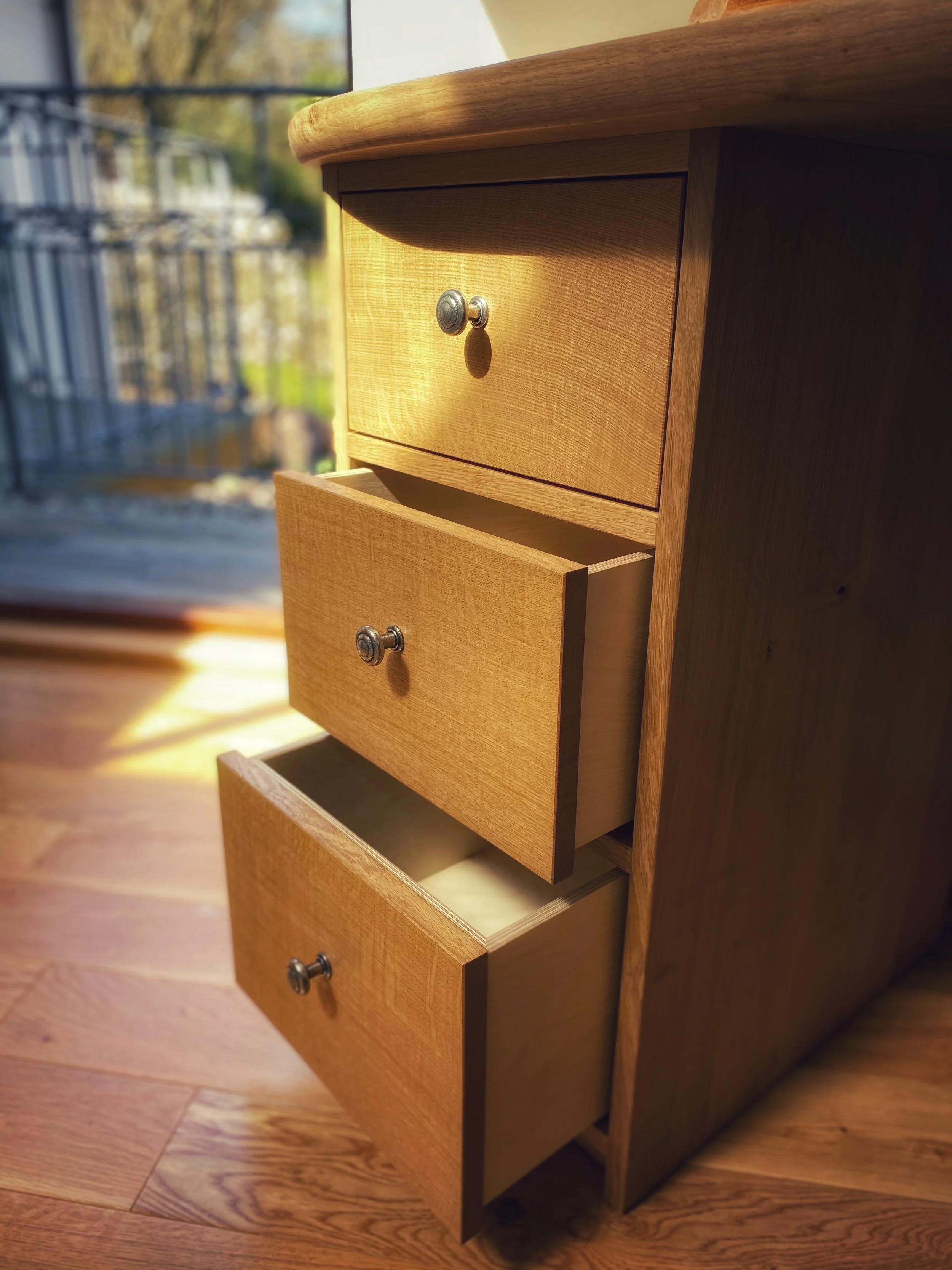 A wooden dresser with three open drawers placed on a hardwood floor near a window, with a balcony and trees visible outside.