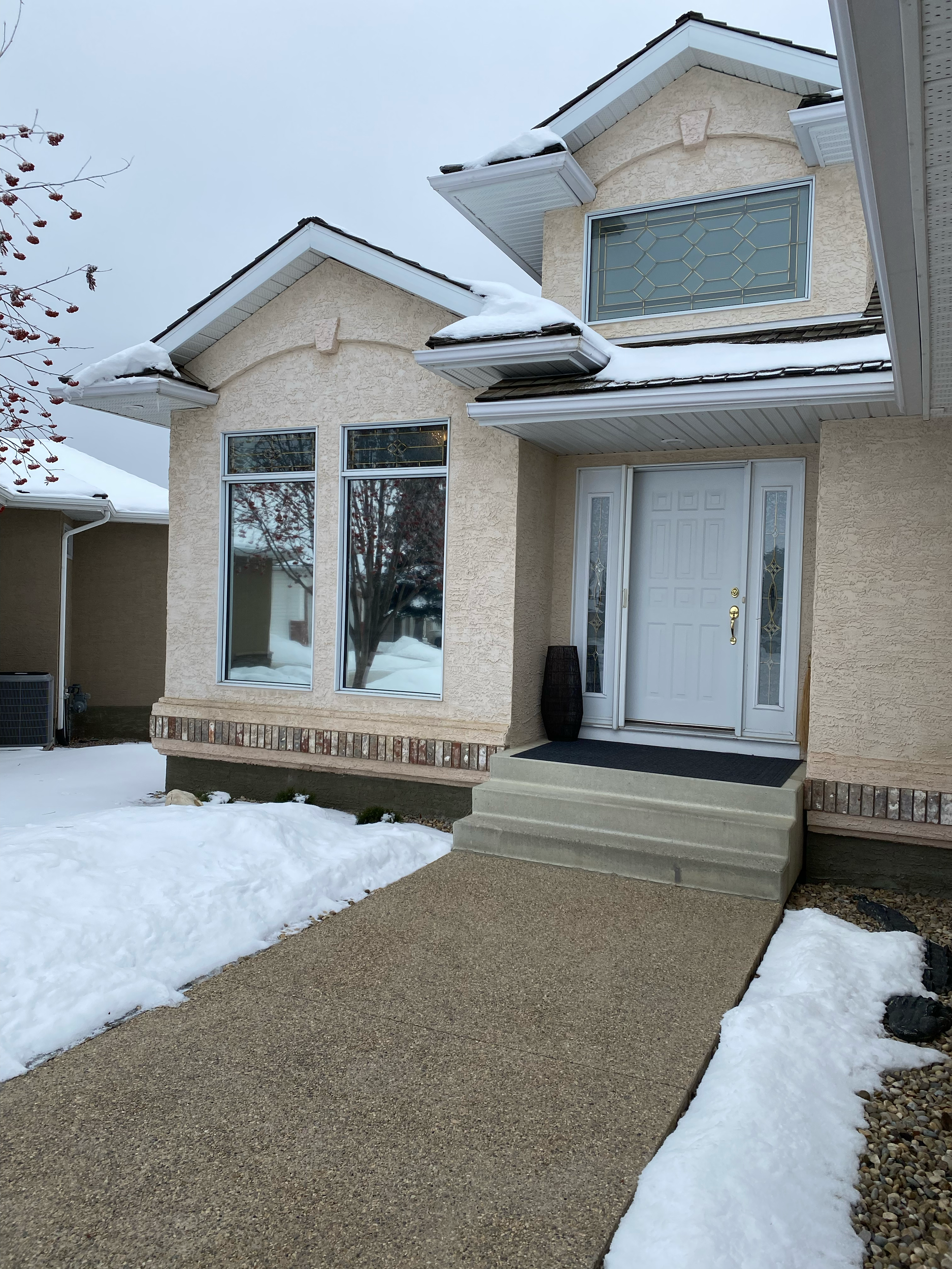 Front entrance of a house with snow-covered steps, a closed white door with decorative glass, and large windows. The house has beige stucco walls and white trim. There is a pile of snow next to the walkway.