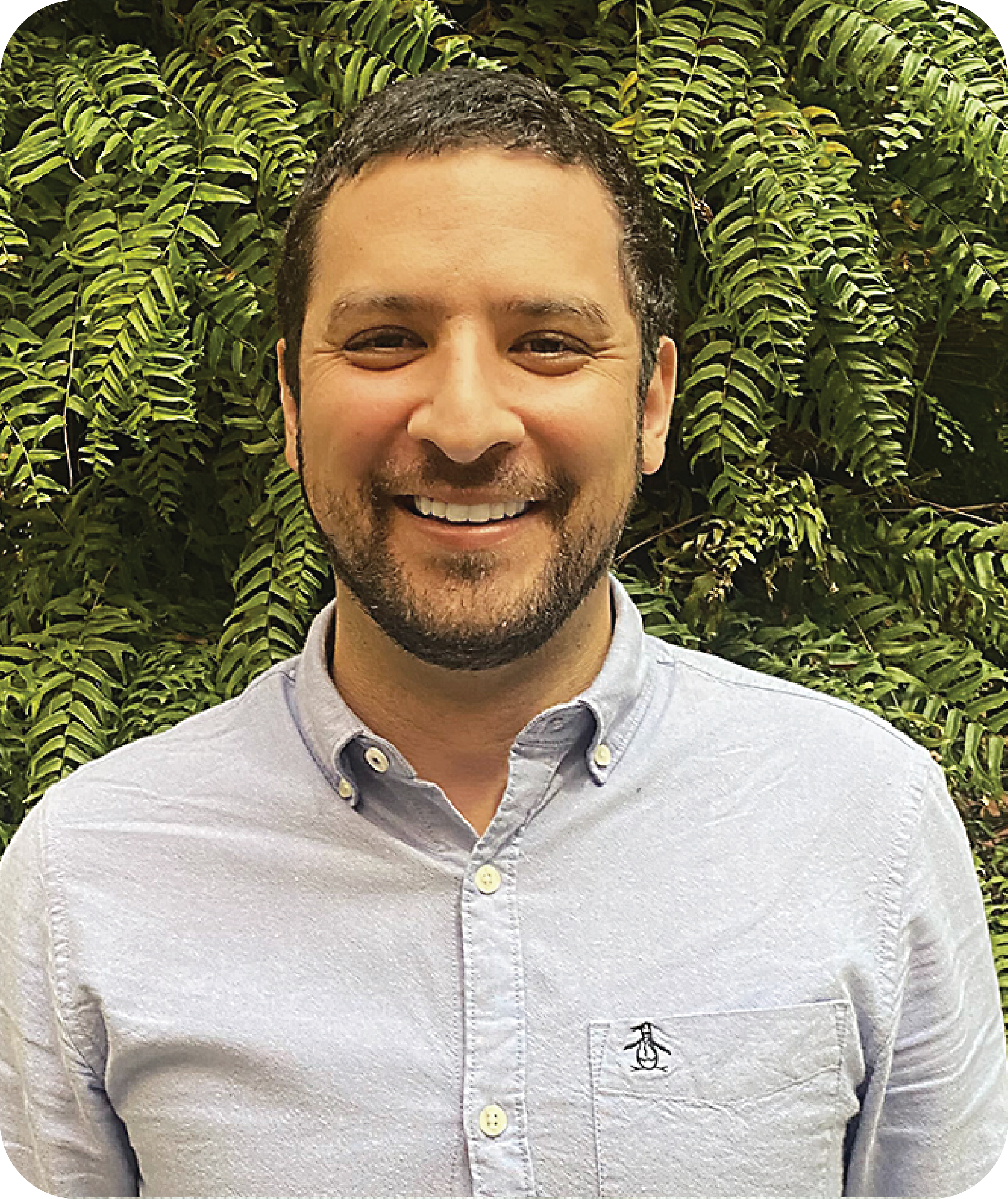 A smiling man with short dark hair and a beard, wearing a light gray button-up shirt with a small penguin logo on the pocket, standing in front of lush green fern leaves.