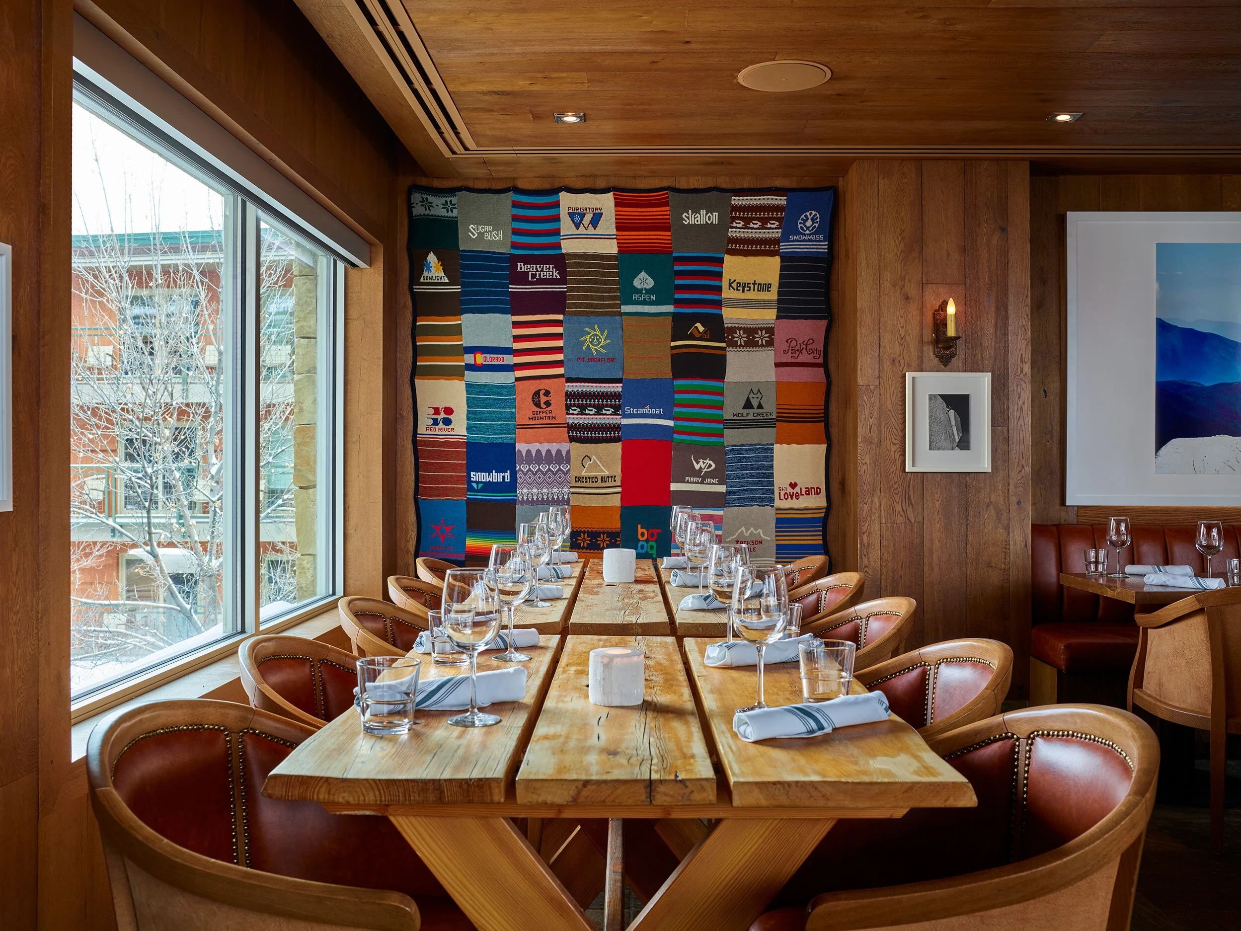 A restaurant dining area with a long wooden table set with wine glasses, water glasses, and napkins. There are leather chairs around the table. A large window on the left shows a snowy landscape outside. The wall has a colorful quilt with various mountain and ski resort patches. The right wall has framed black-and-white art and a sconce light.