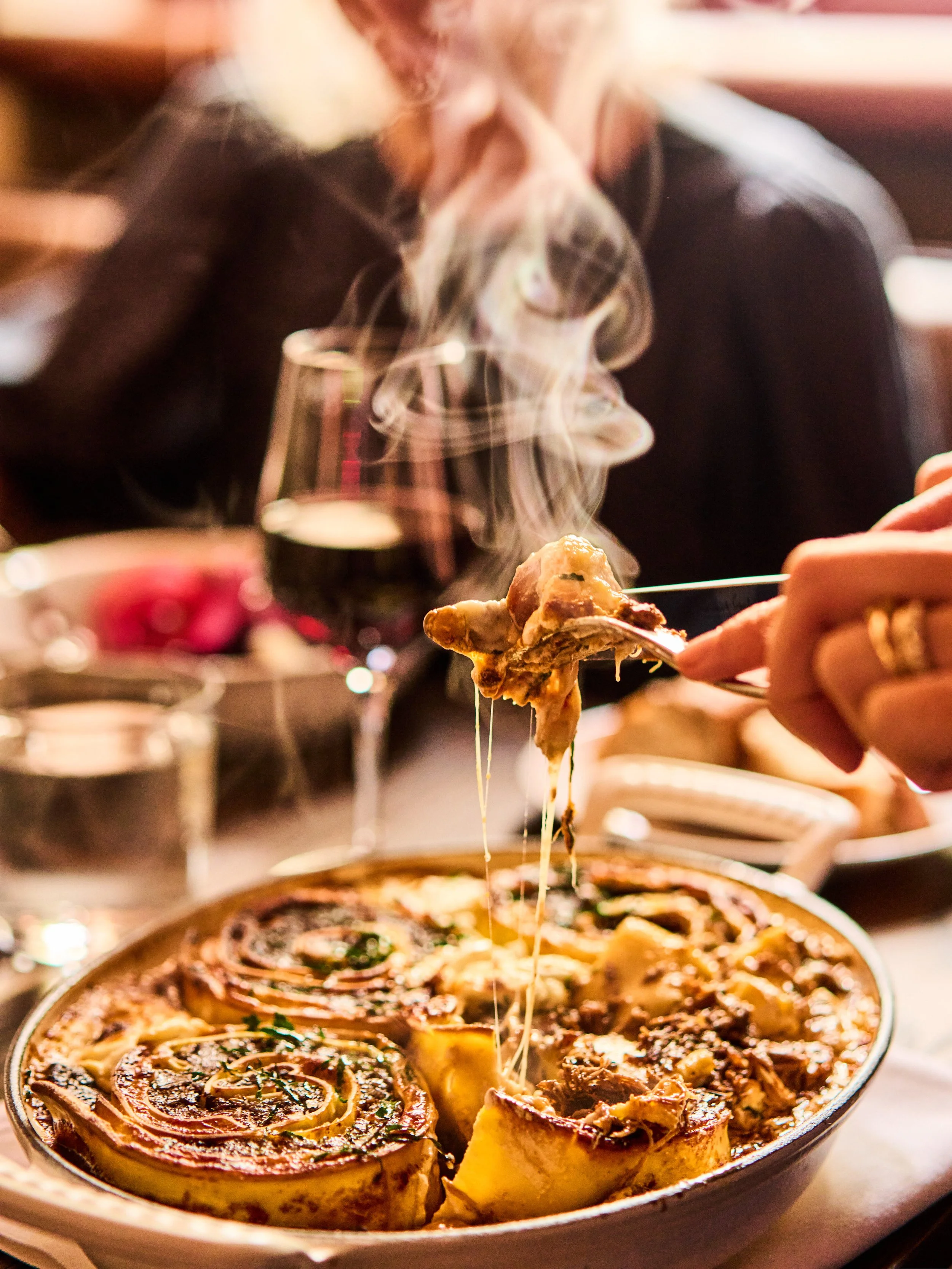 A person is holding a fork with a piece of food over a dish of baked eggplant rollatini topped with cheese and herbs, with more eggplant rollatini inside a baking dish.