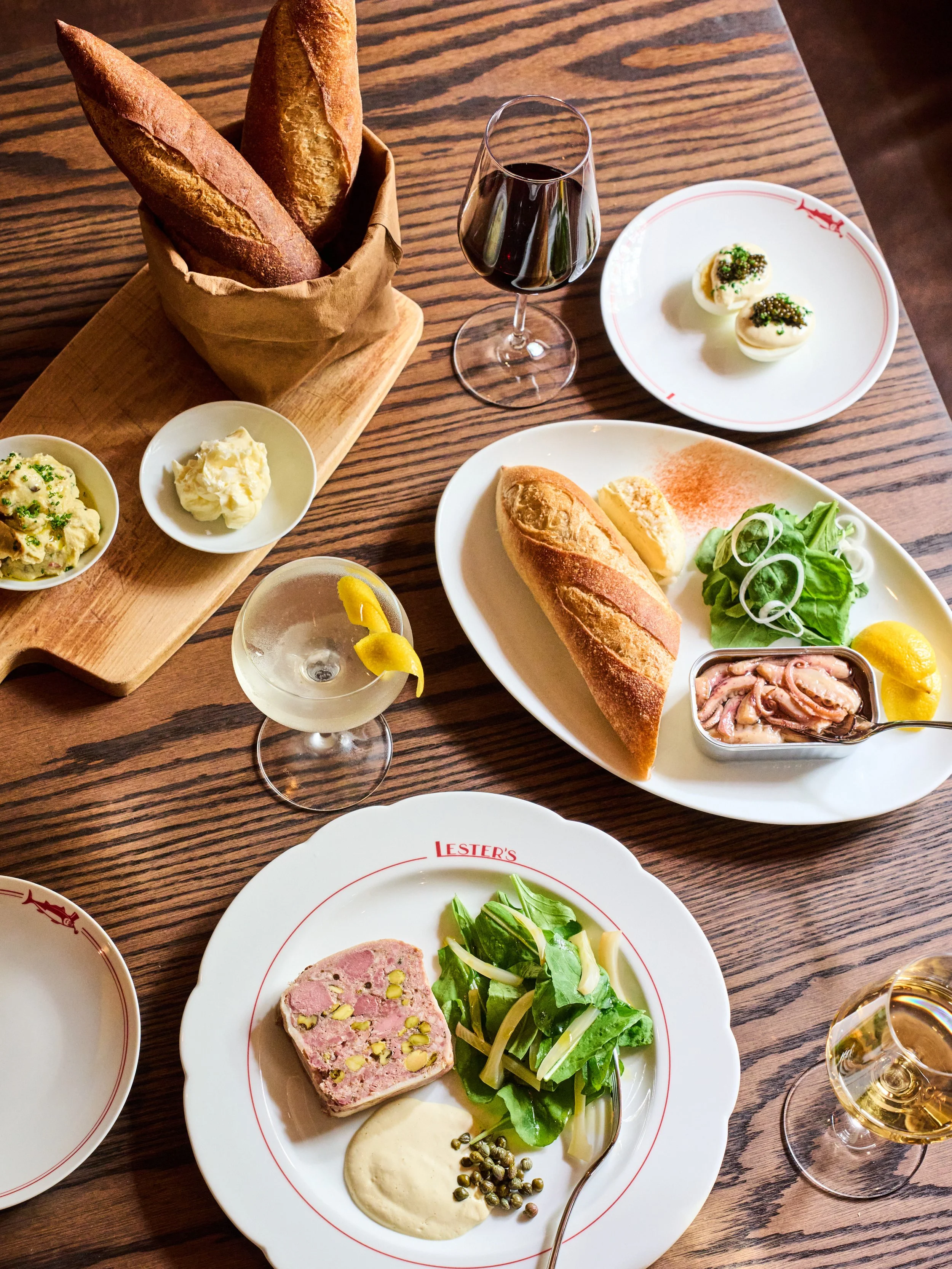 A table set with various foods including baguette bread in a basket, a glass of red wine, deviled eggs sprinkled with caviar, a plate with a baguette, a slice of pâté garnished with pistachios, greens, and a dollop of cream, along with a smaller dish