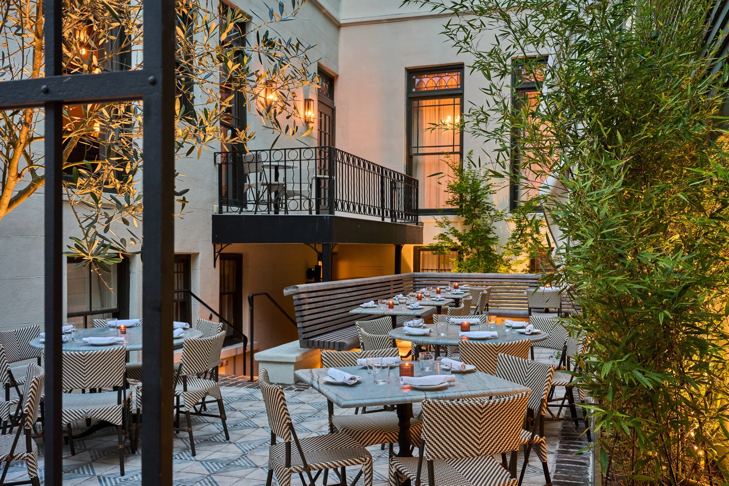 An outdoor dining area with tables set with napkins, glasses, and candles, surrounded by greenery and featuring a building with balconies and large windows, lit by warm wall lights.
