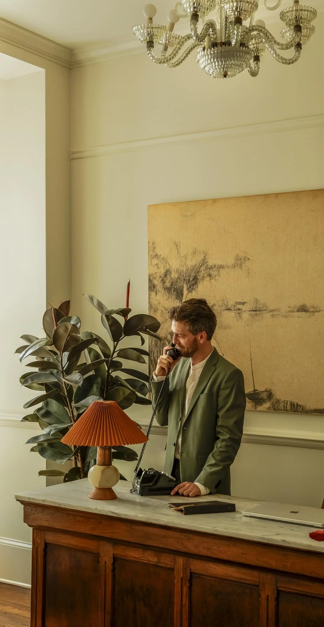 A man in a green blazer and white shirt talking on a vintage rotary telephone inside a room with elegant decor. There is a large potted plant, a table lamp with a pleated shade, a marble-topped wooden desk, a closed laptop, and a framed landscape painting on the wall.
