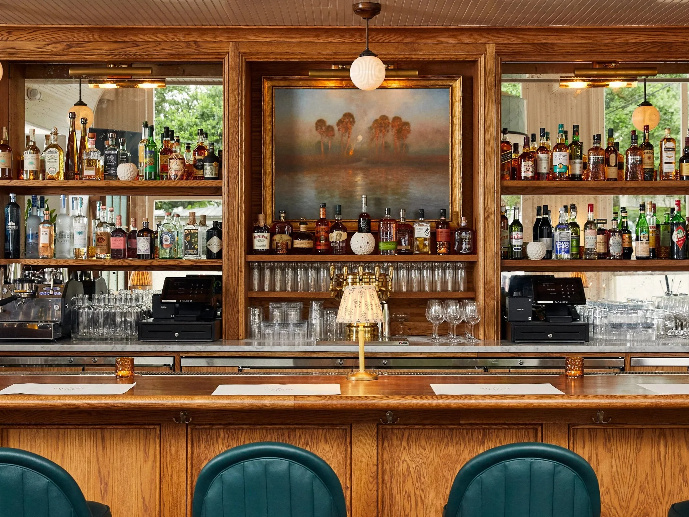 Interior of a bar with wooden shelves stocked with various liquor bottles. A painting of trees and water is centered above the bar, with a small lamp and glassware below. Green chairs are visible in the foreground.