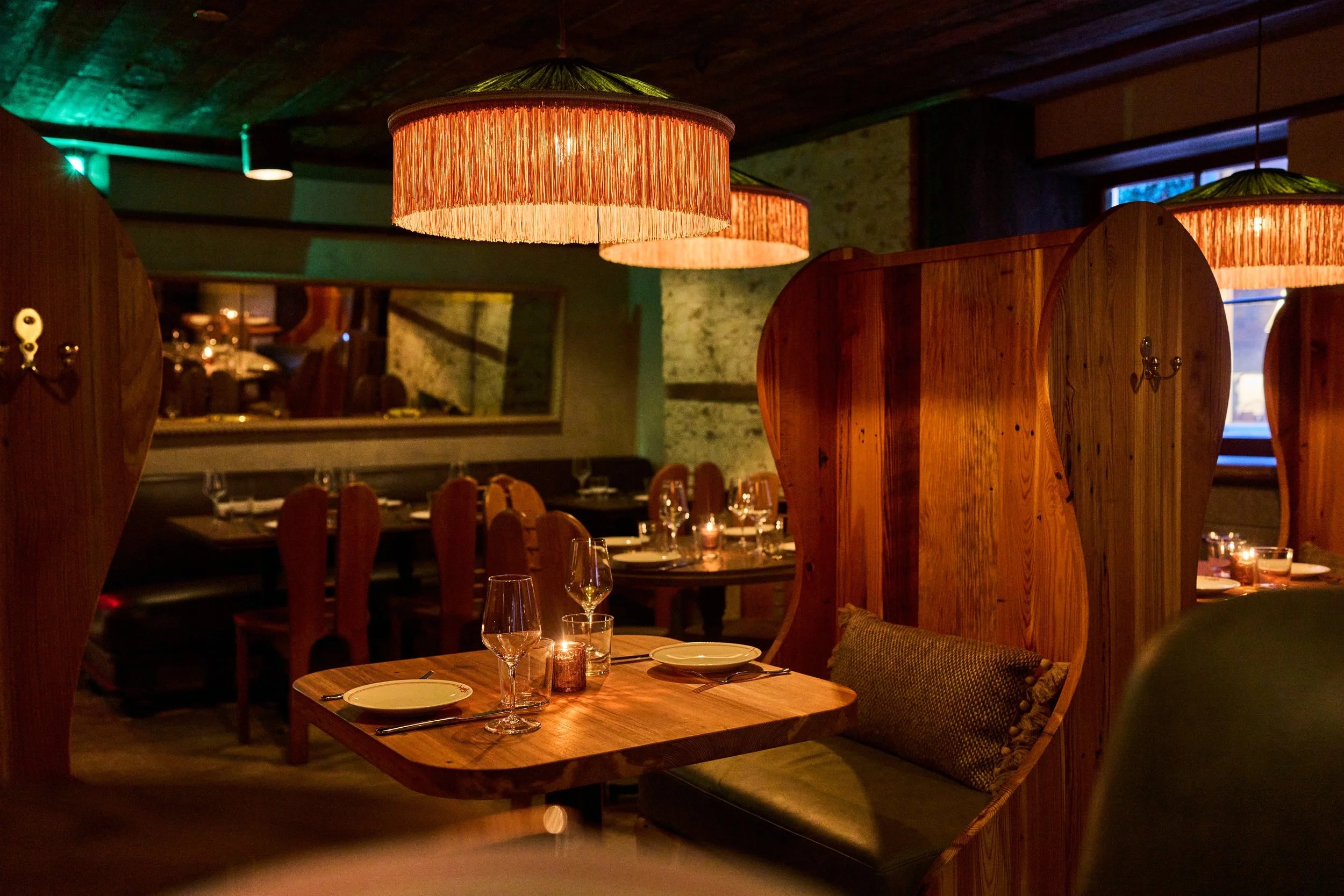 Interior of a cozy restaurant with wooden partitions, tables set with wine glasses, plates, and candles, warm lighting from fringed pendant lamps, and a mirror on the wall enhancing the intimate atmosphere.