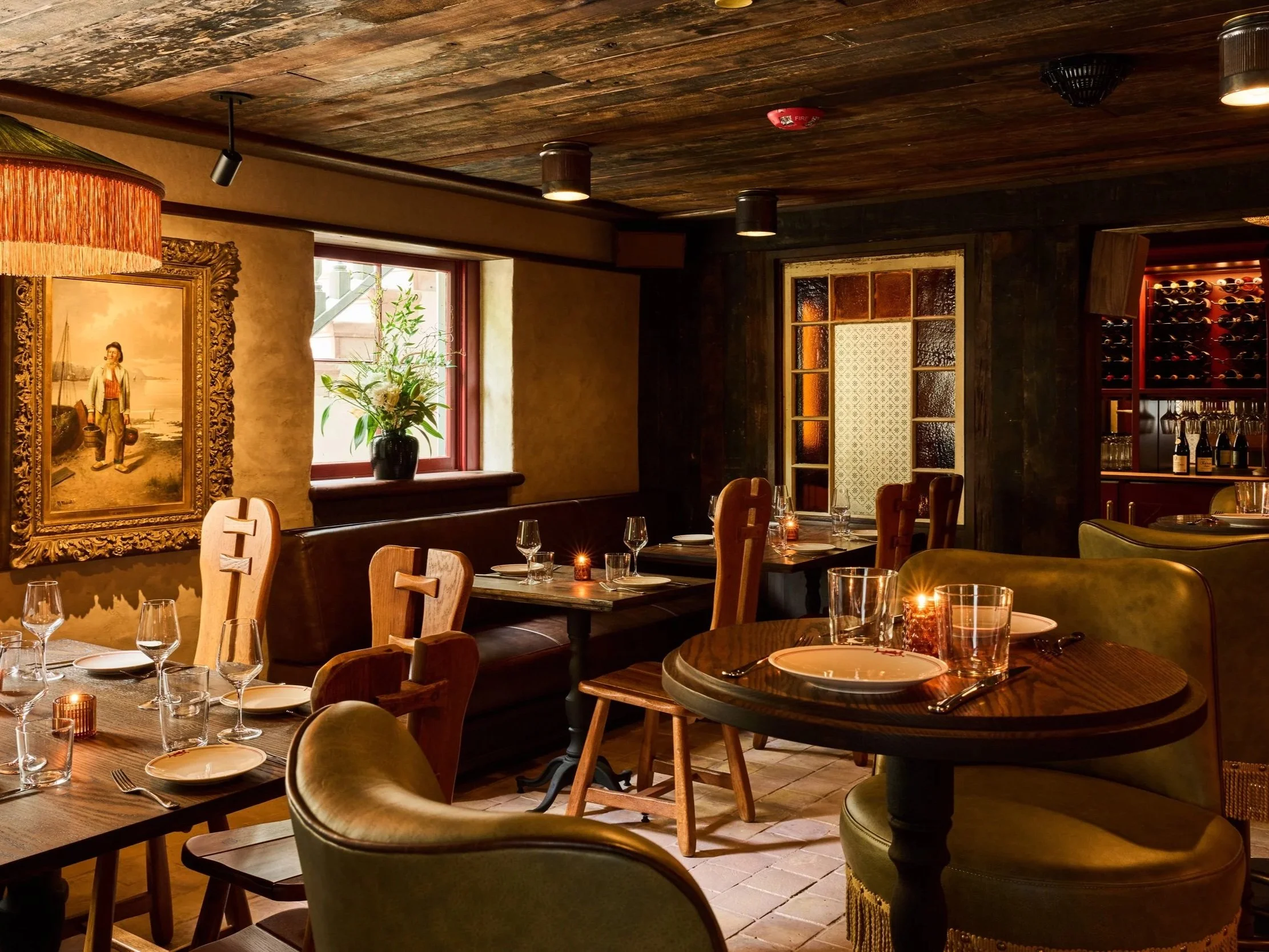 A cozy restaurant interior with wooden tables and chairs, set with glasses, plates, and candles for dining, featuring warm lighting, a window with greenery outside, artwork on the wall, and a wine display in the background.