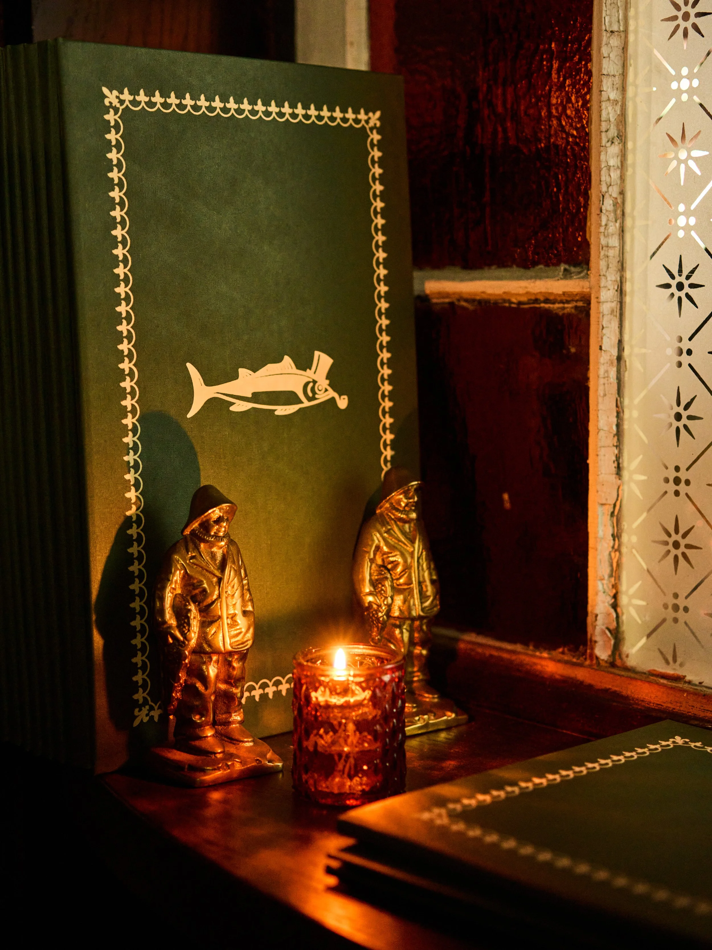 Devotional altar with two gold-colored statuettes, a burning candle in a glass holder, a green cloth with a white embroidered border, and a window with decorative metalwork in the background.
