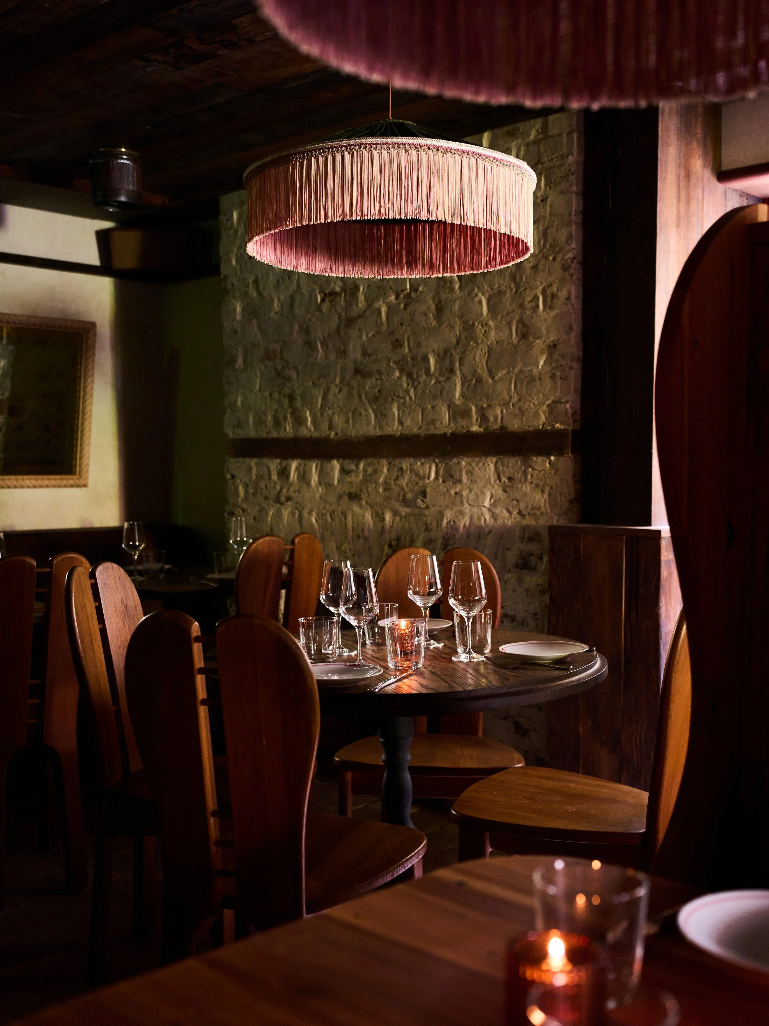 A cozy restaurant with wooden tables and chairs, a rustic brick wall, and warm lighting from candles and a large pink fringed lampshade hanging from the ceiling.