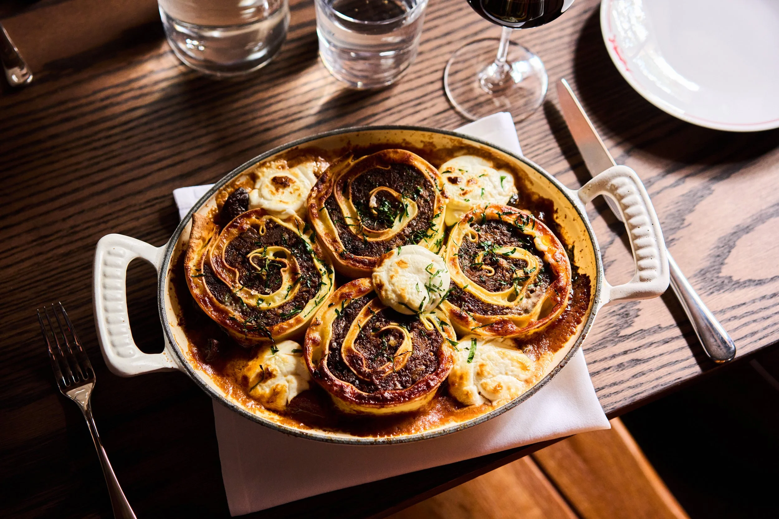 A baked dish of lasagna with visible layers of pasta, sauce, cheese, and herbs in a white oval dish, on a wooden table with a wine glass, drinking water glasses, and plates nearby.