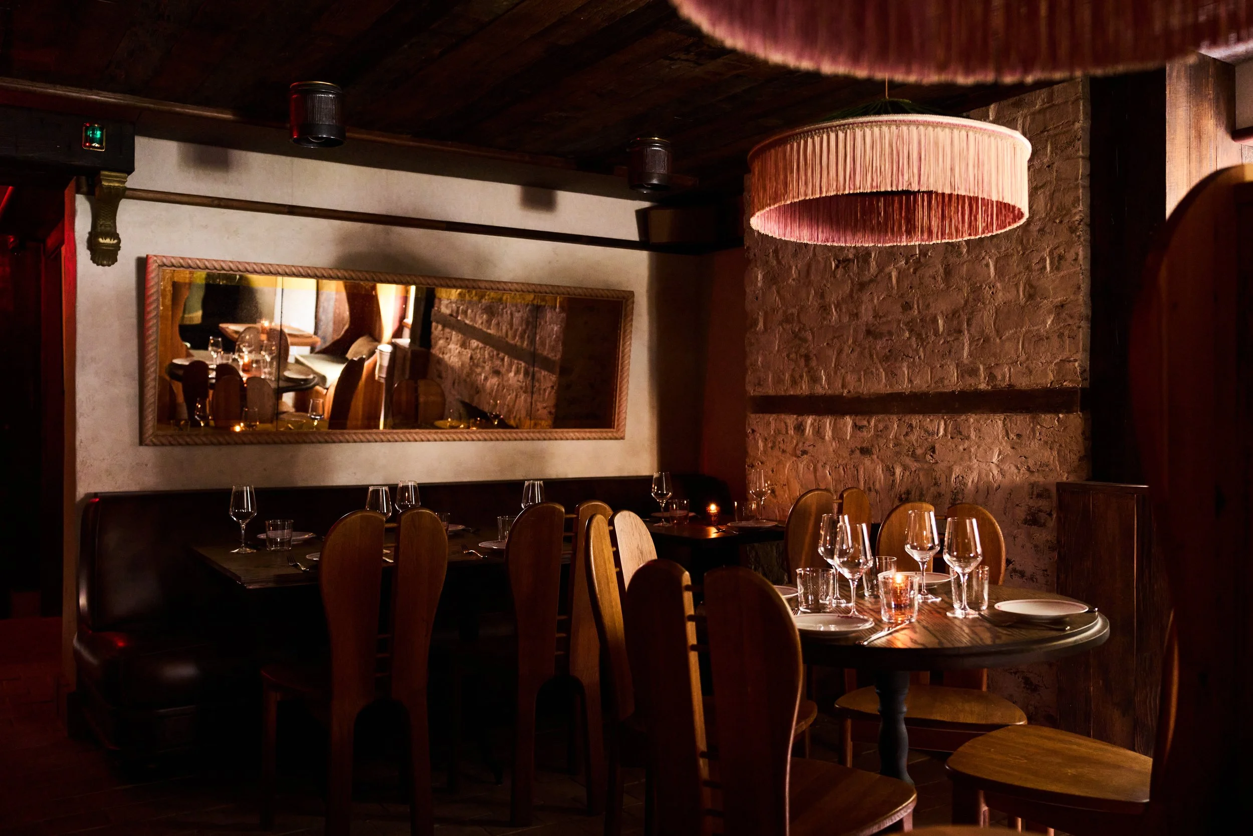 Interior of a dimly lit restaurant with wooden chairs and tables set with wine glasses and plates, a large pink fringed hanging lamp, brick walls, and a tall mirror reflecting the dining area.