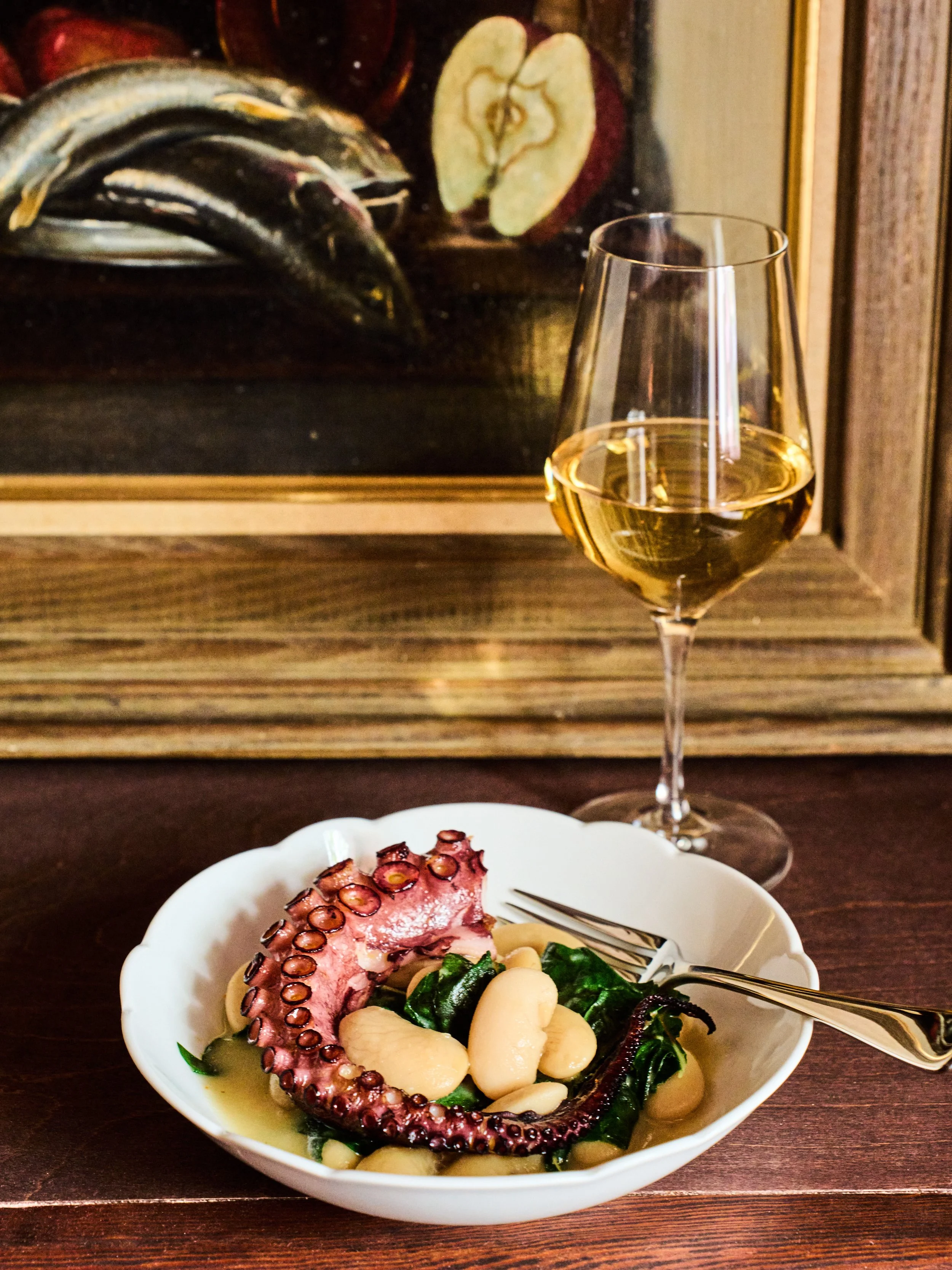 A white bowl containing cooked octopus tentacle, white beans, and leafy greens. In the background, a glass of white wine and a painting of apples are visible.