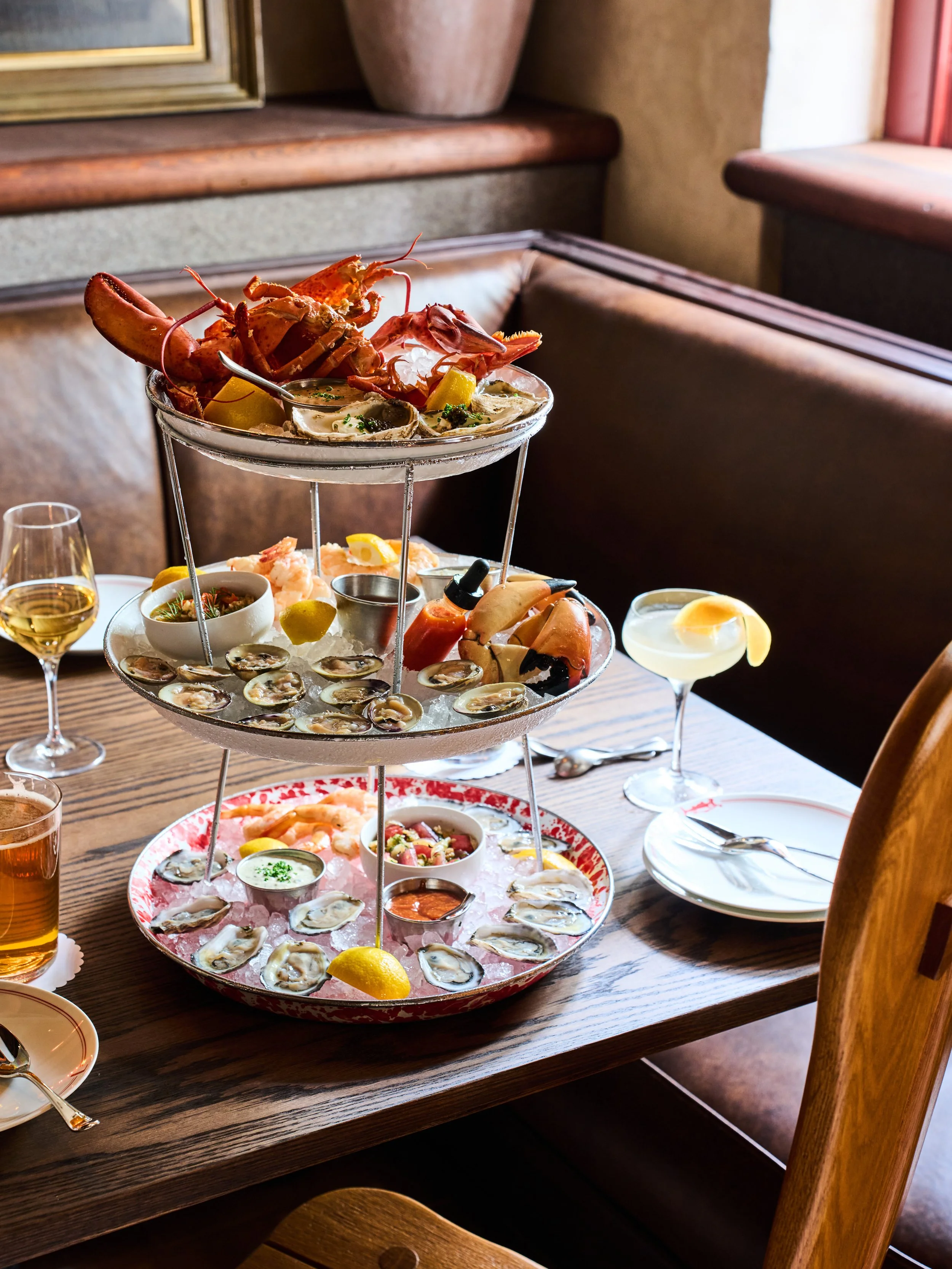 A multi-tiered seafood platter on a wooden table next to a glass of white wine, a cocktail, and other dishes in a cozy restaurant setting.
