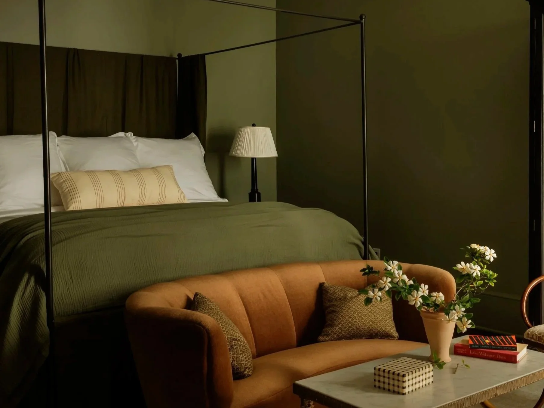 A cozy bedroom with a made bed, a lamp, an orange sofa with patterned pillows, a vase with white flowers, and books on a marble coffee table.