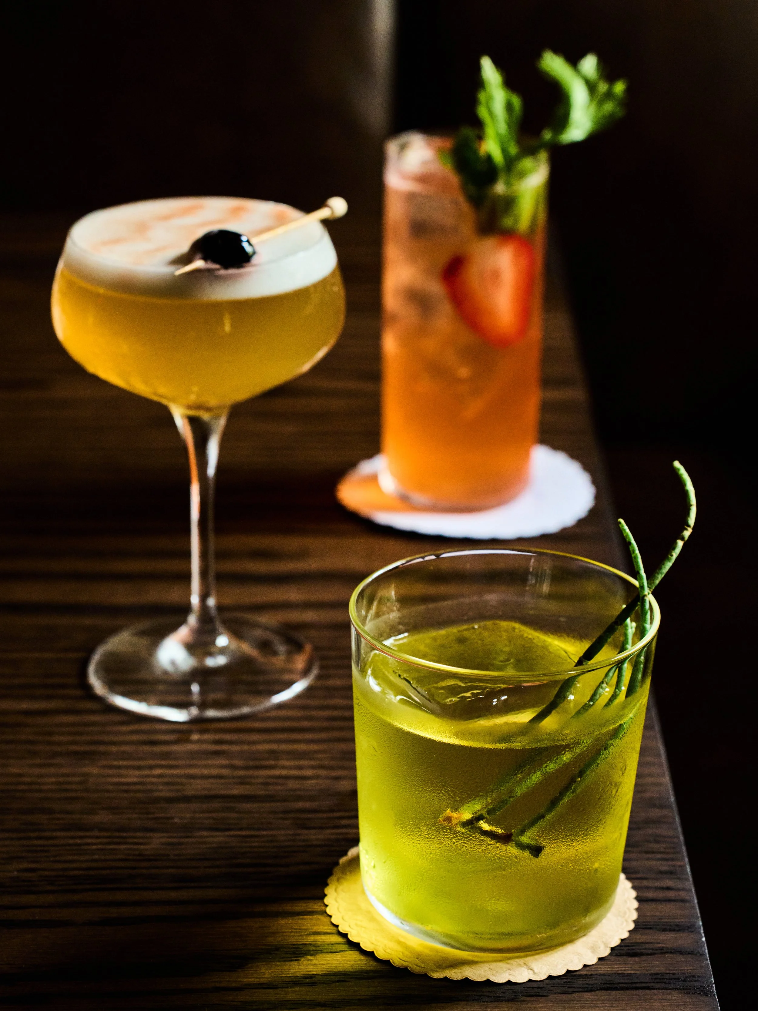 A yellow cocktail with ice and cherry garnish on a wooden table, with a clear glass containing a green drink with mint leaves and a sprig of herbs in the foreground, and a tall glass with a strawberry and mint in the background.