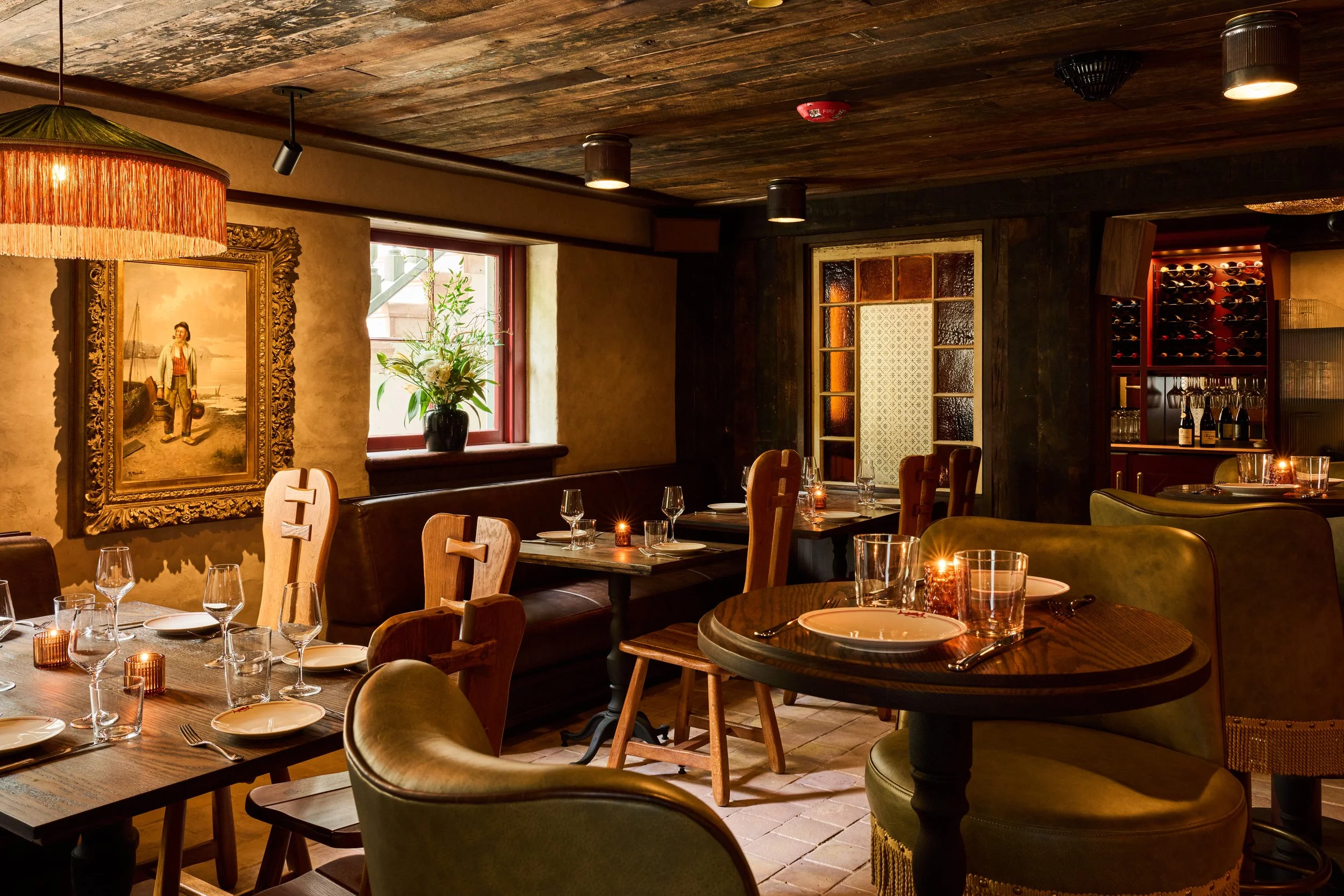 A cozy restaurant interior with wooden tables set for dining, surrounded by green upholstered chairs. There is a large painting of a woman on the wall, a window with a vase of flowers near it, and warm lighting creating an inviting ambiance.
