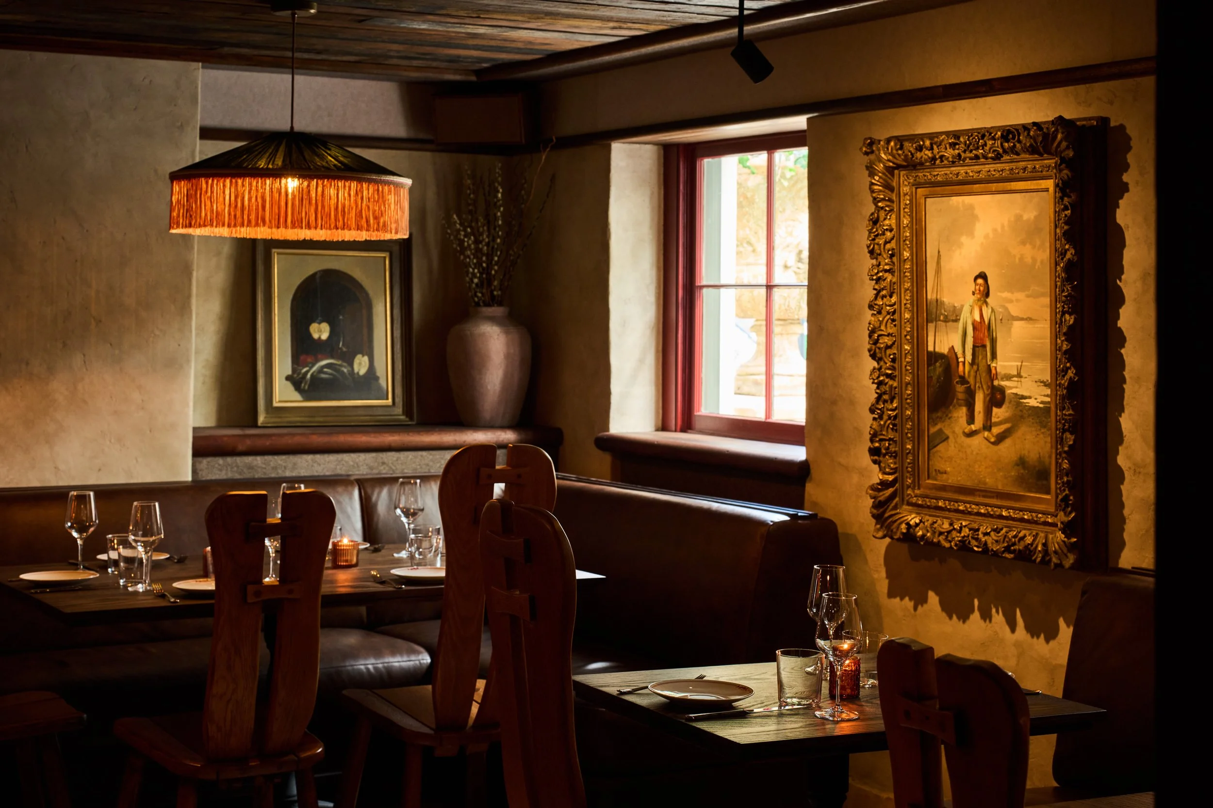 Interior of a cozy restaurant with wooden furniture, framed artwork, decorative vase, and warm lighting from a hanging lamp, near a window with natural light.