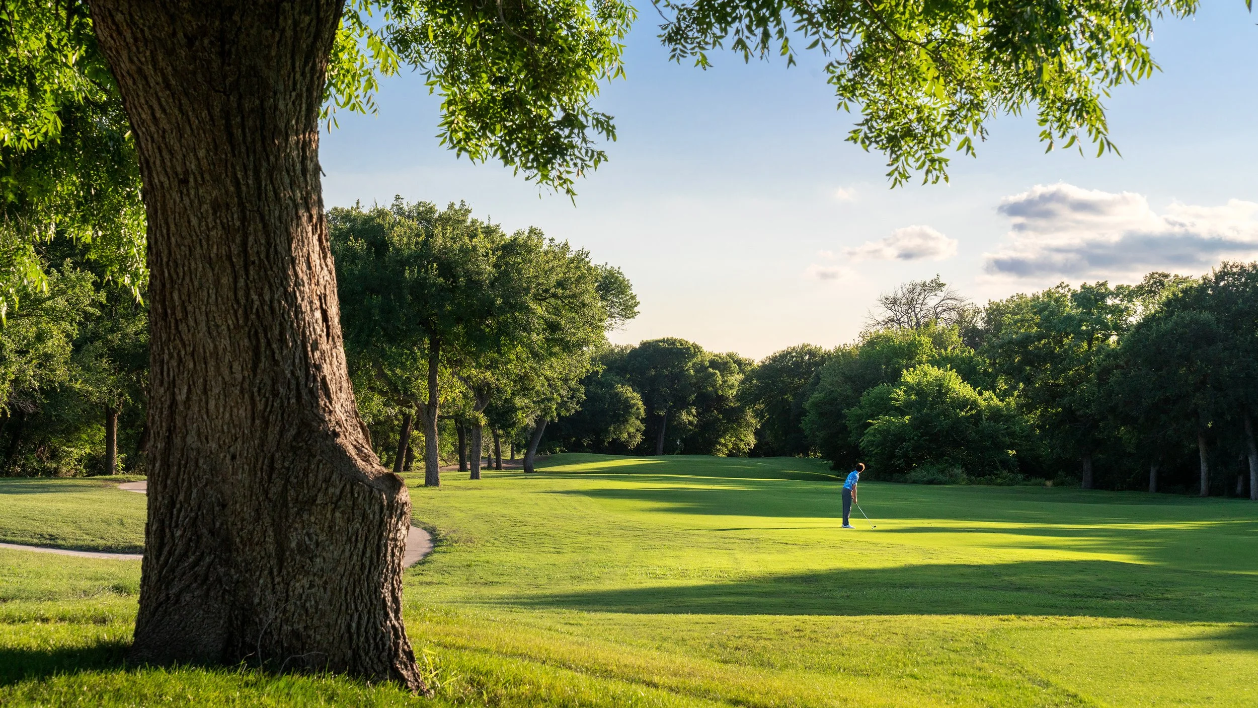 A person playing golf on a green golf course surrounded by trees under a blue sky with scattered clouds.