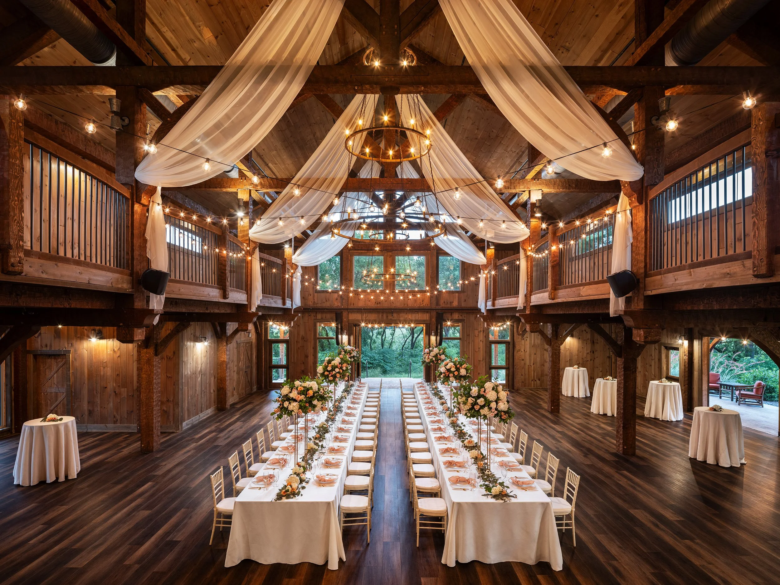 A rustic wedding venue with a long banquet table decorated with bouquets of pink and white flowers, set in a barn with wooden beams, draped white fabric, and string lights hanging from the ceiling.