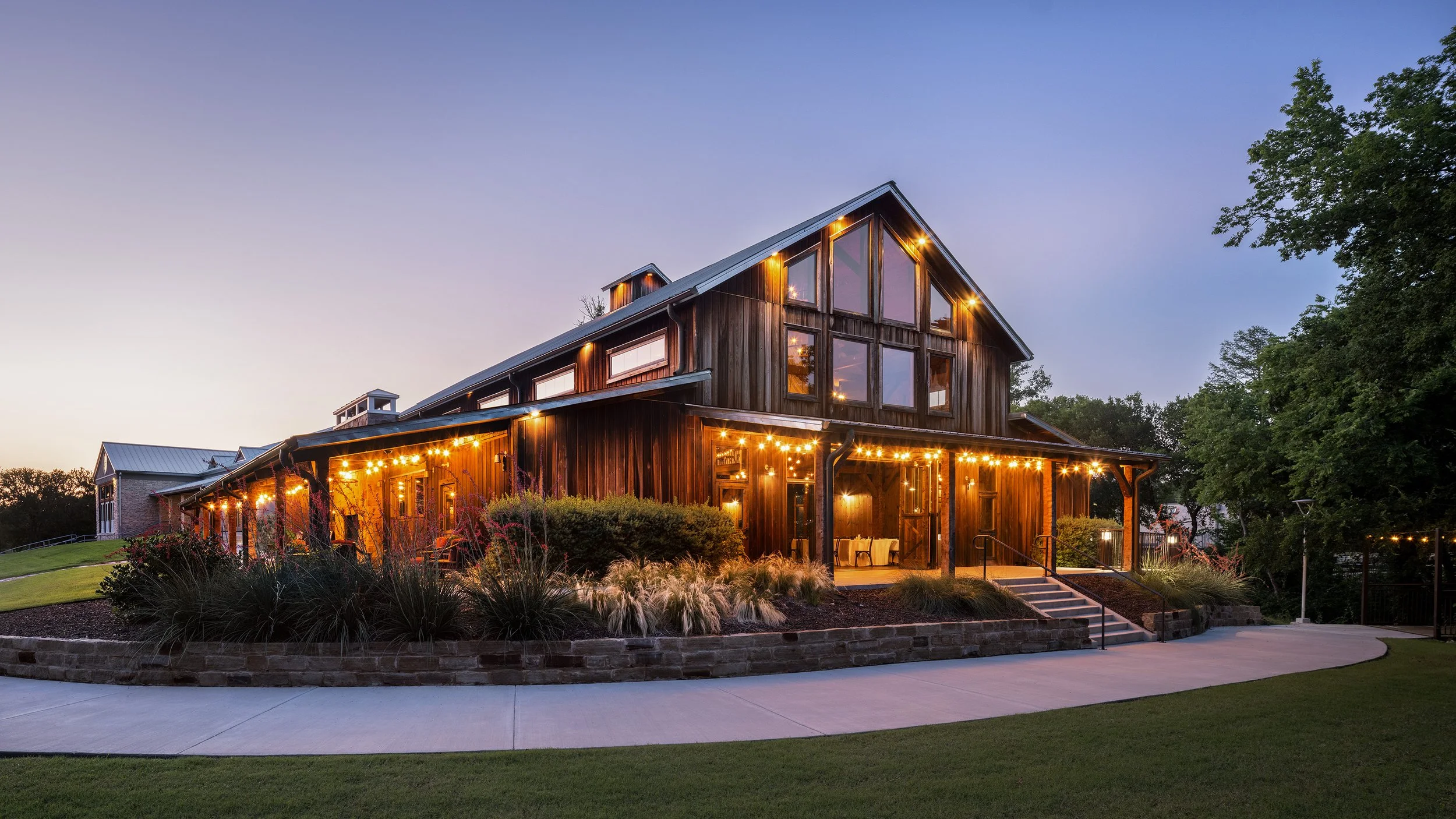 A rustic wooden house with large, angled windows and string lights illuminating the porch area at dusk, surrounded by manicured lawn and trees in the background.
