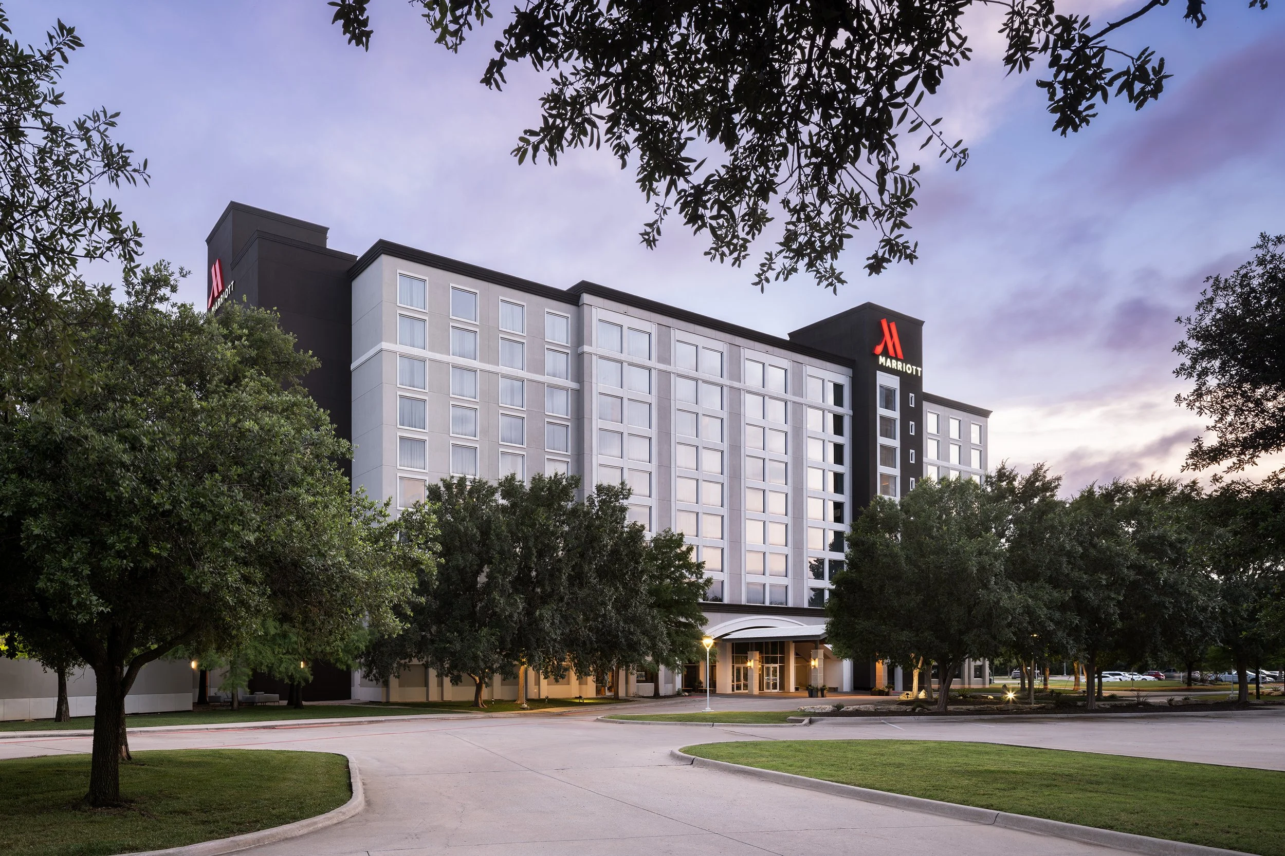A mid-rise hotel building with the Marriott logo on the top, surrounded by trees and a parking lot, during dusk or early evening.