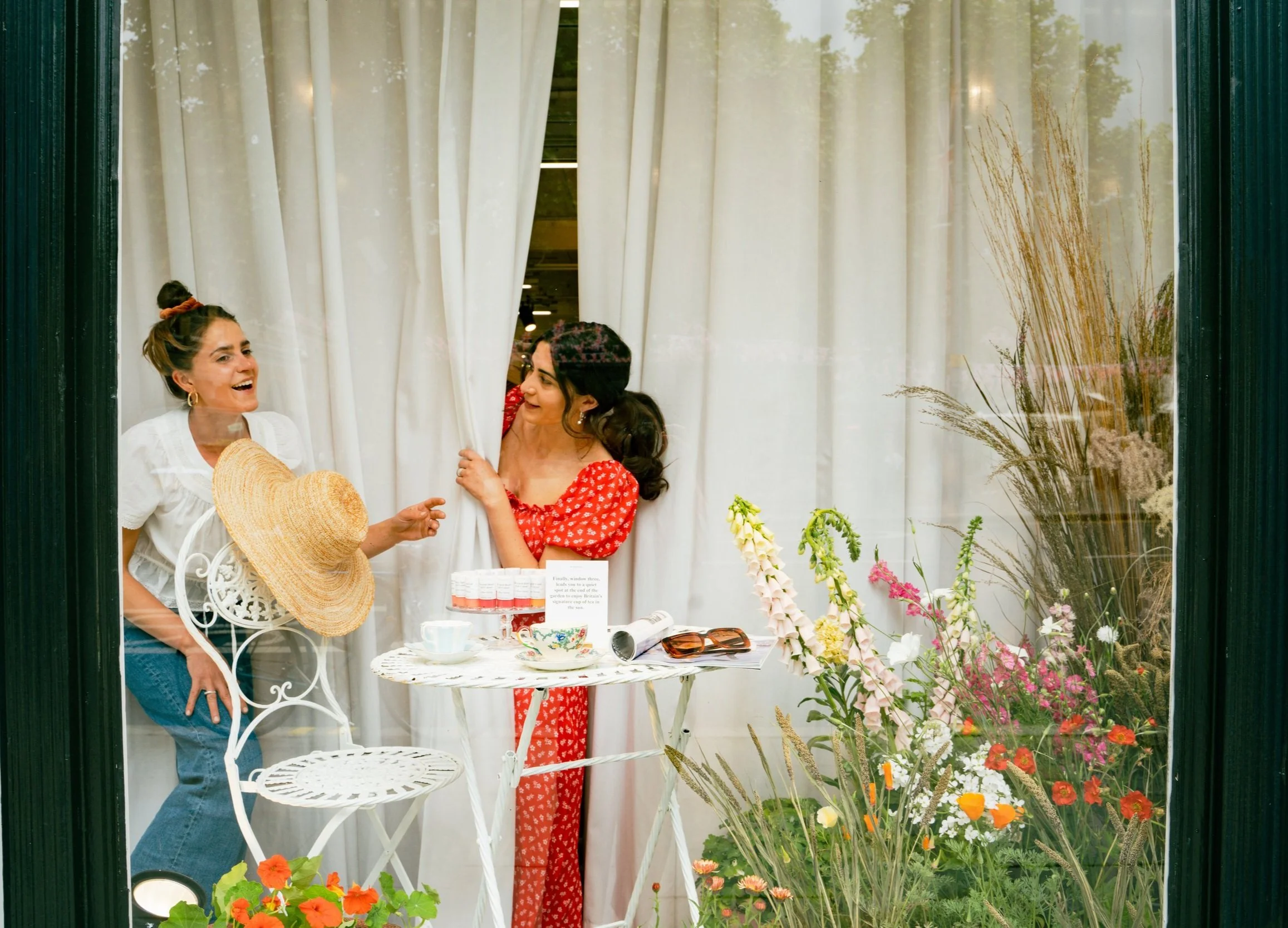 Two women are inside a floral shop, smiling and talking. One woman is holding a large straw hat and wearing a white top and blue jeans, while the other woman, in a red dress, is peeking from behind a curtain. The shop has flowers on the right side an