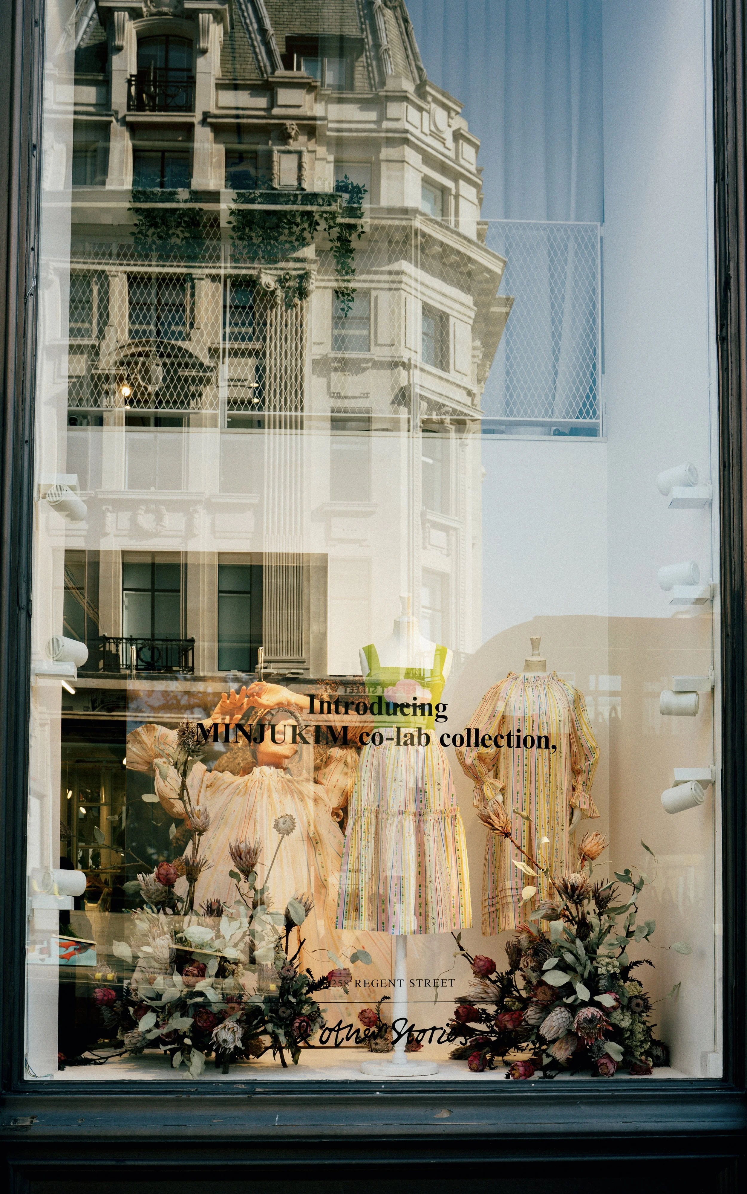 Store window display showcasing mannequins dressed in colorful, striped dresses with floral arrangements at the bottom. Reflects buildings and blue sky outside.