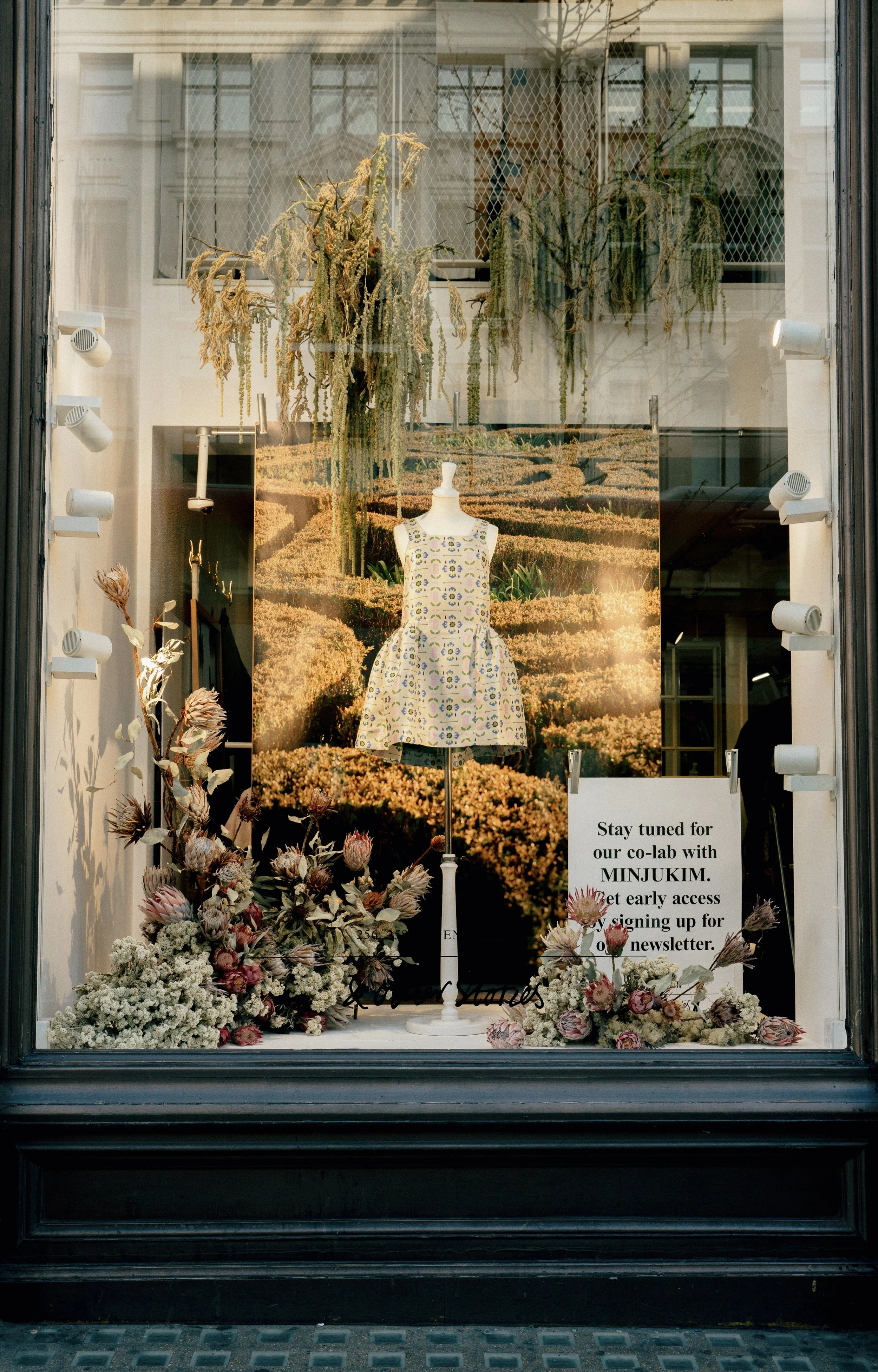 Store window display with a mannequin wearing a patterned dress, surrounded by dried flowers and a sign that invites customers to join a co-lab event with MINJUKIM and sign up for a newsletter. The background features a large landscape image.