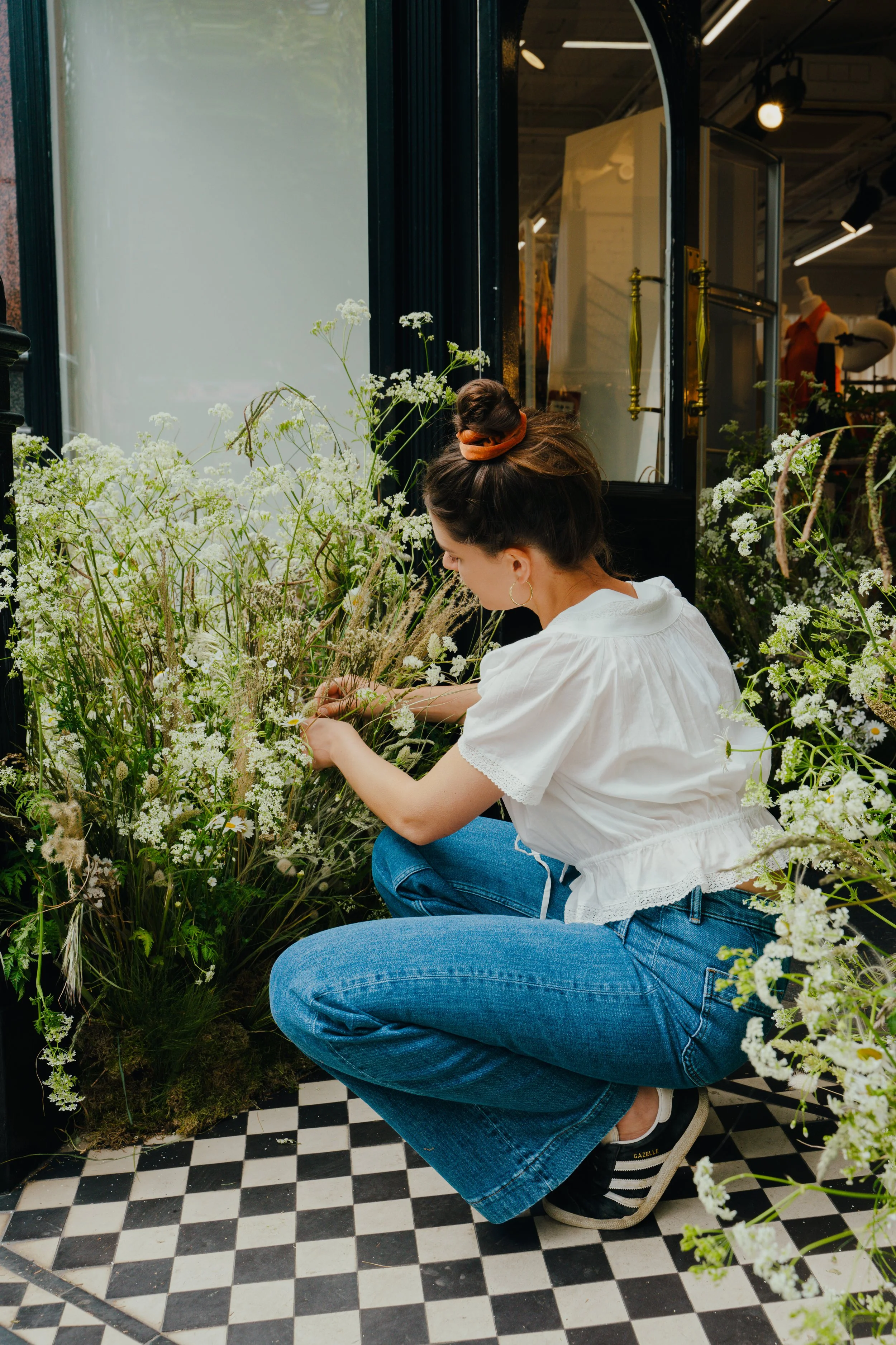 Woman crouching near white flowers inside a shop with black and white checkered floor.