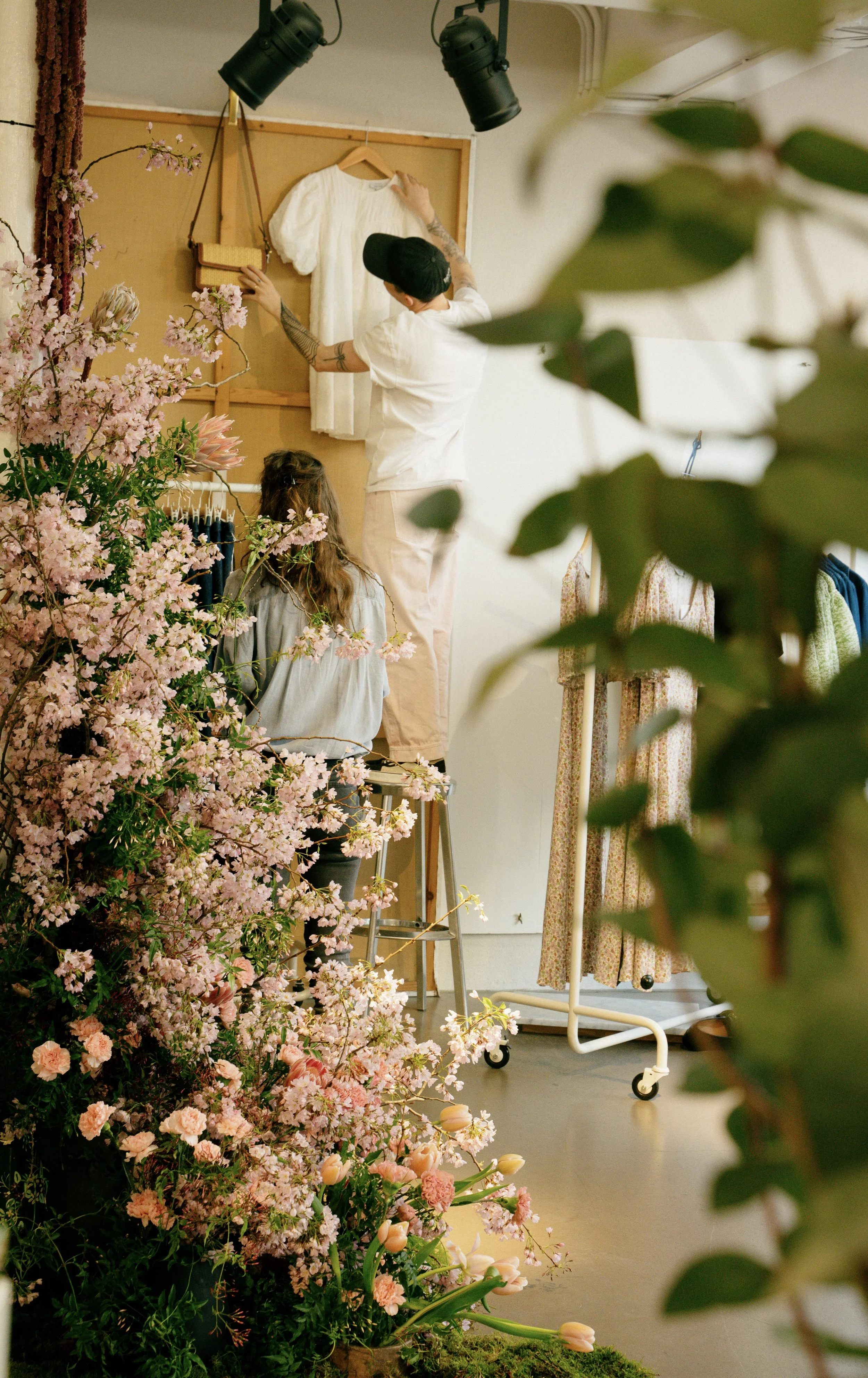 People arranging clothes in a boutique with pink flowers in the foreground.