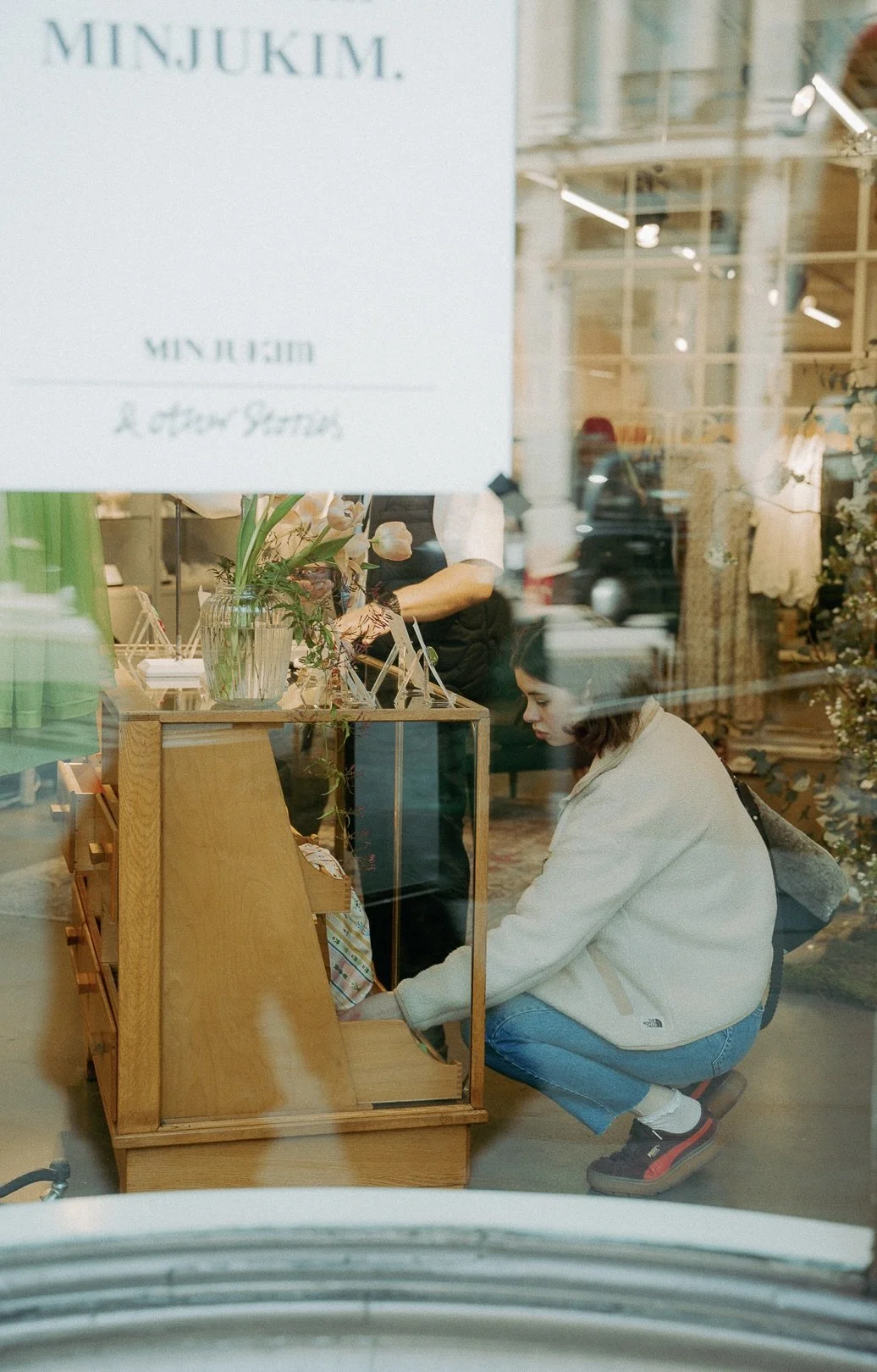A person crouching down and browsing inside a wooden display case with glass sides, inside a flower shop. The shop has a sign that reads 'MINJUKIM.' through the glass, which reflects the street outside.