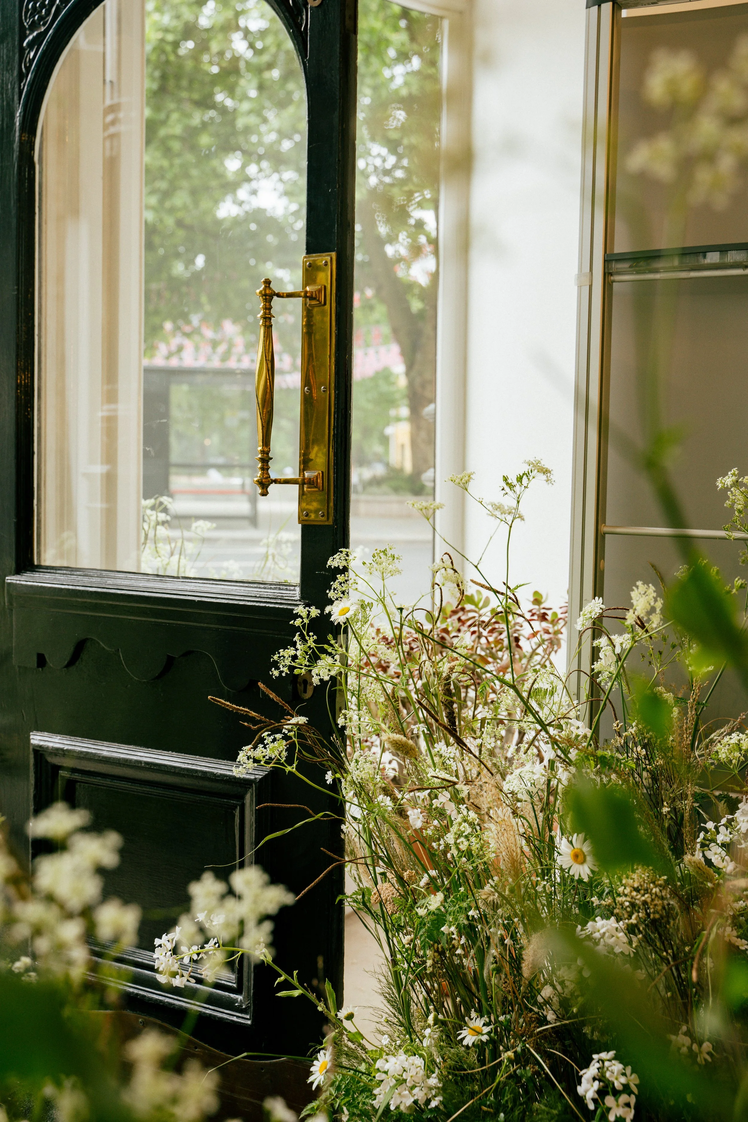 Open black door with brass handle leading outside, with a view of trees and a sidewalk; inside, a bouquet of white and green wildflowers on the floor.