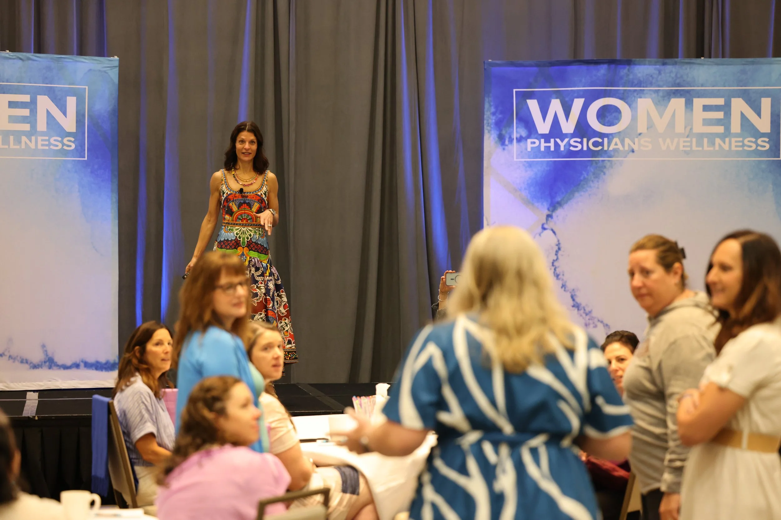 A woman stands on a stage at a conference or event, with a sign behind her reading 'WOMEN PHYSICIANS WELLNESS'. She is speaking to an audience of women seated at tables, some of whom are looking towards her or taking photos.