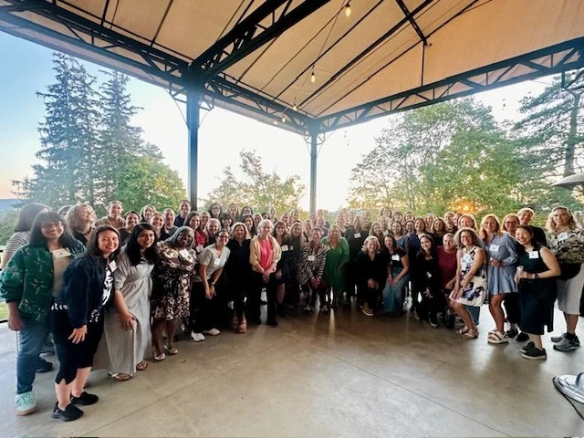 Group of diverse women gathered outdoors under a covered pavilion, smiling for a photo with trees and sunset in the background.