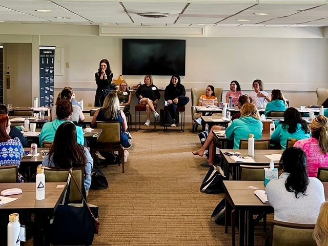 A woman is speaking to a group of people seated in a conference room, with four panelists on a stage and a large screen behind them.