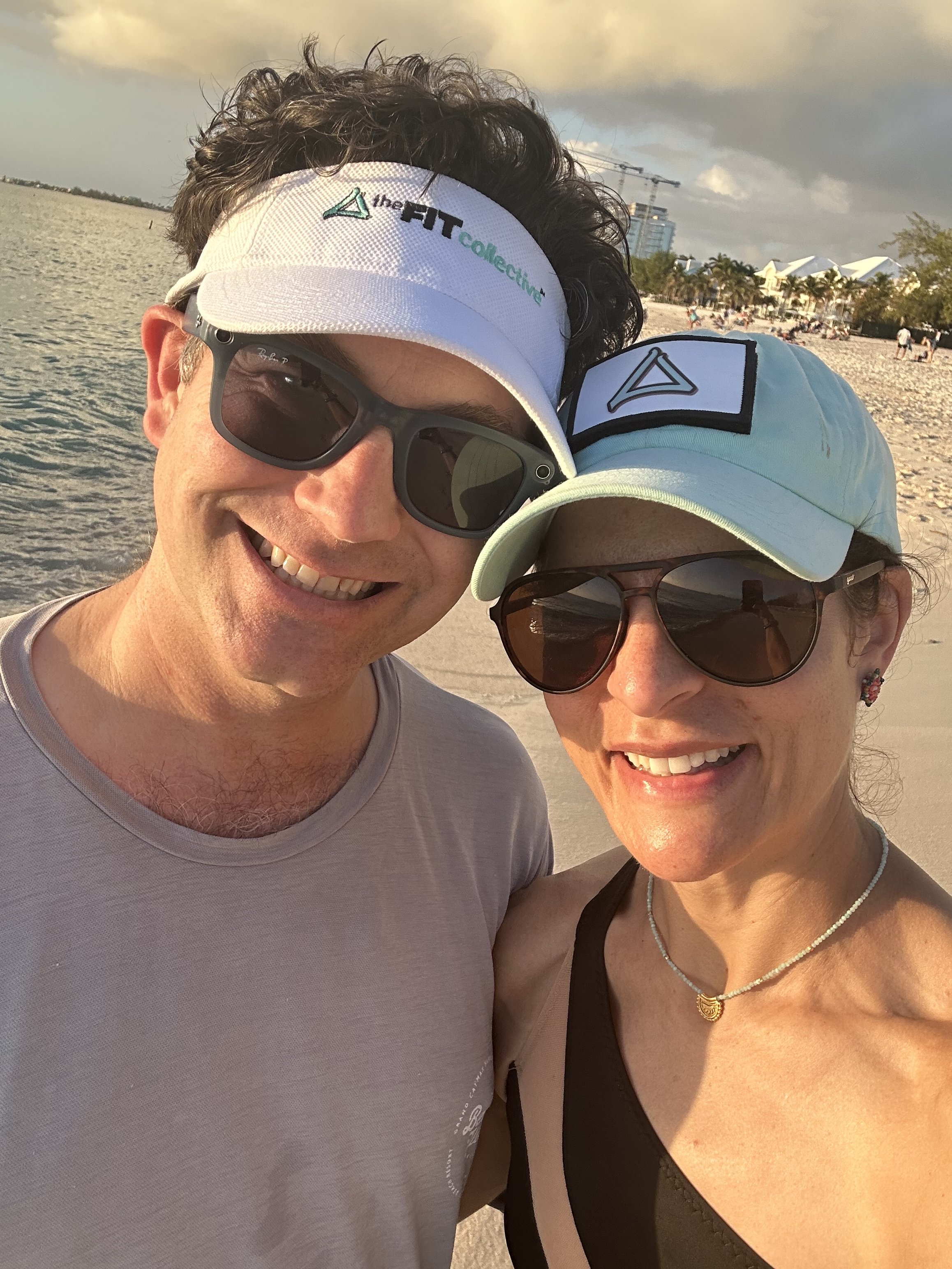 A smiling man and woman taking a selfie on the beach during sunset. They are wearing sunglasses and hats, with a beach and some buildings visible in the background.