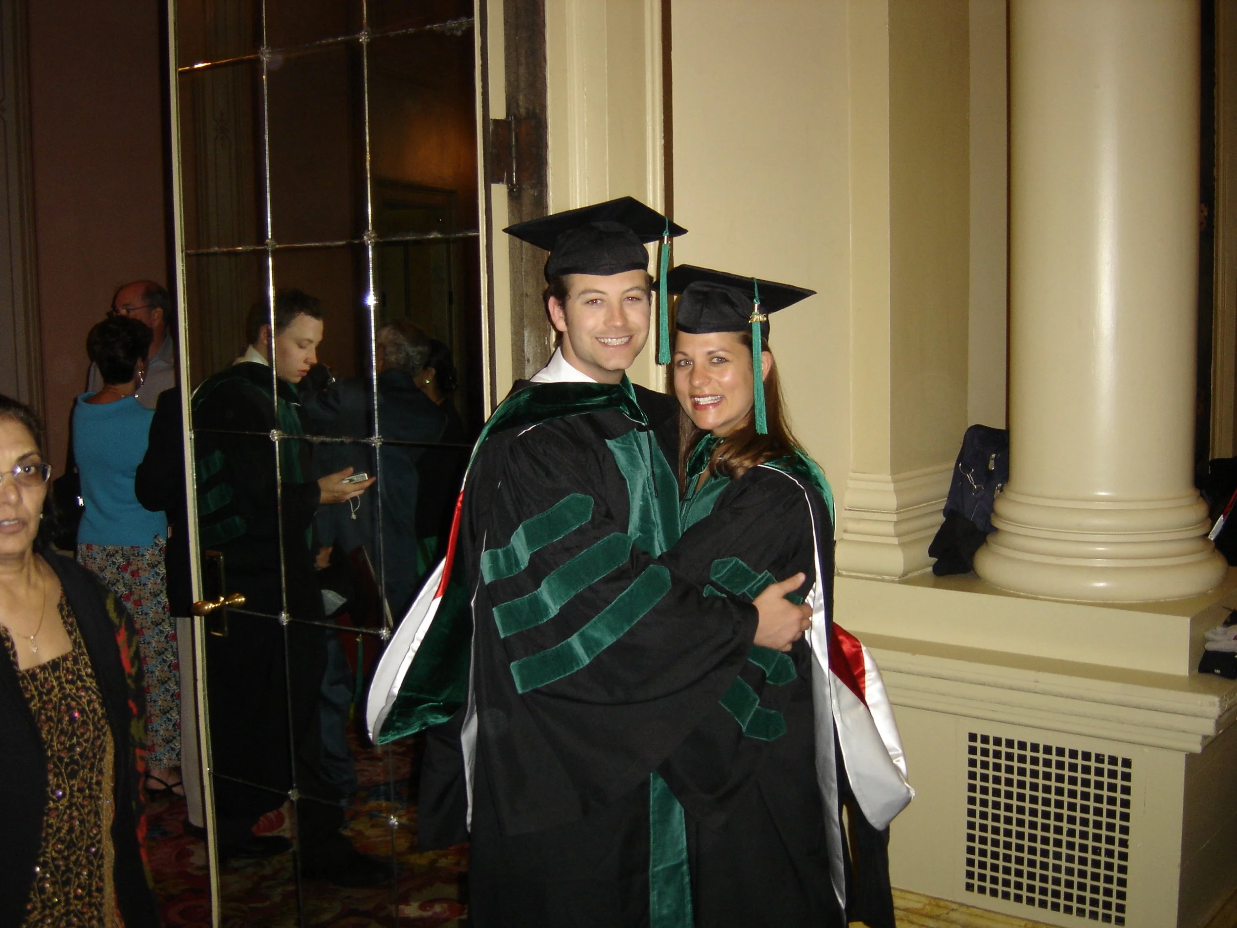 Two graduates in green and black robes and mortarboards hugging at a graduation ceremony indoors with mirror and column in the background.