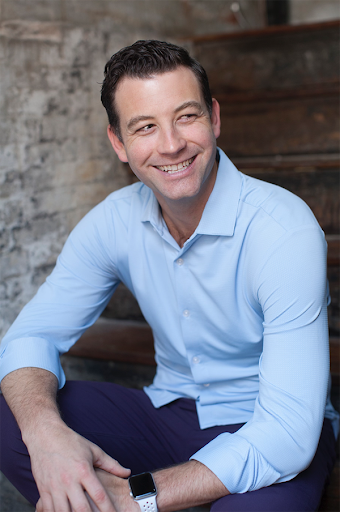 Smiling man with dark hair, wearing a light blue shirt, sitting outdoors against a brick and wooden background.