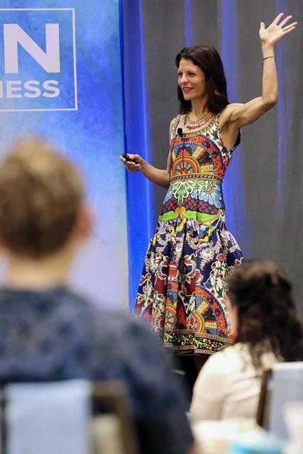 A woman in a colorful patterned dress is giving a presentation on stage, smiling and waving to the audience, with a blue backdrop and a partially visible sign.