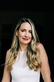 A woman with long wavy hair smiling at the camera, wearing a white sleeveless top.