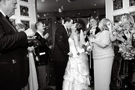 A bride and groom holding hands at their wedding reception, surrounded by guests, some holding flowers and drinks, in a warmly decorated indoor setting.