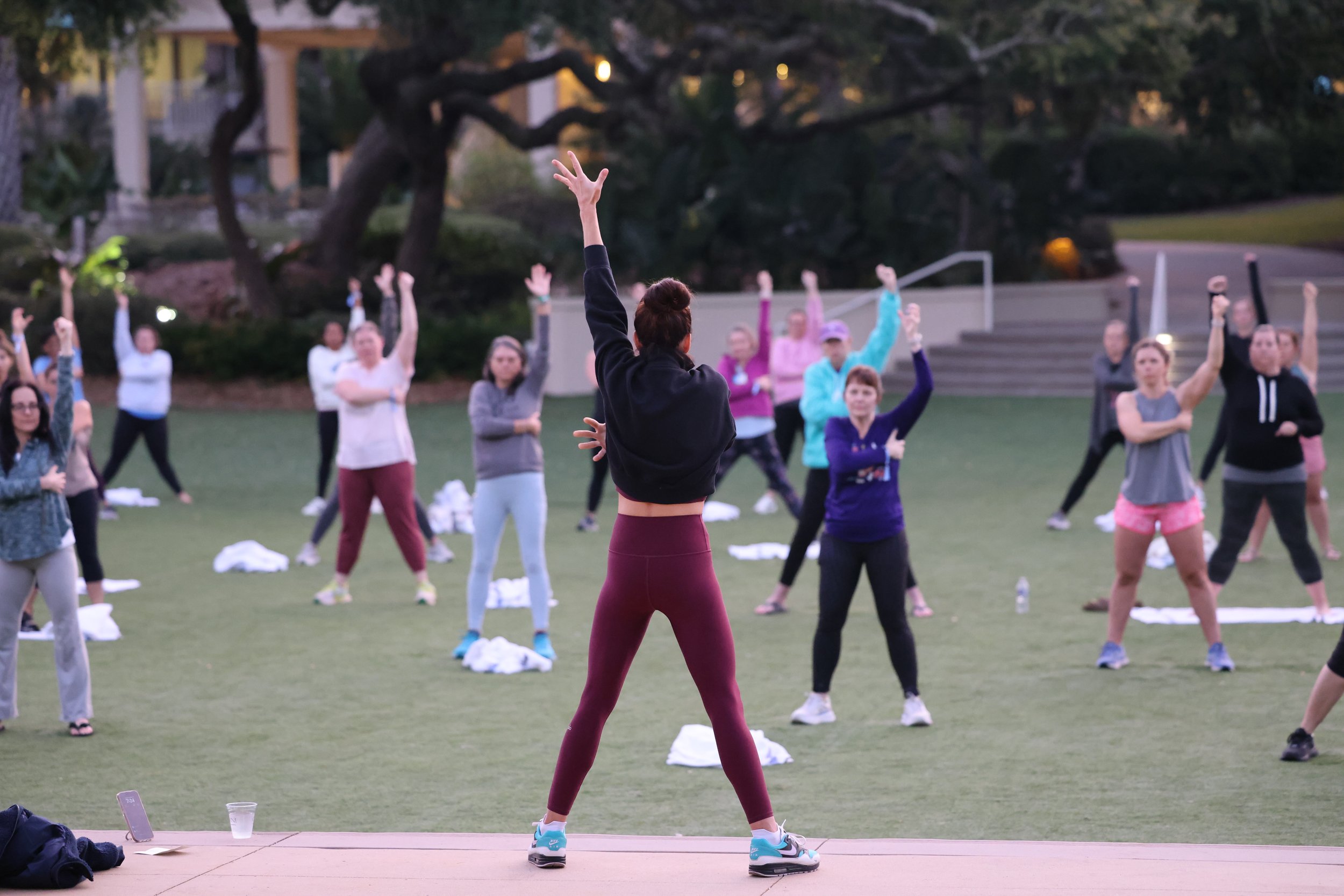 Instructor leading outdoor group fitness class with participants stretching and following movements on a grassy area.