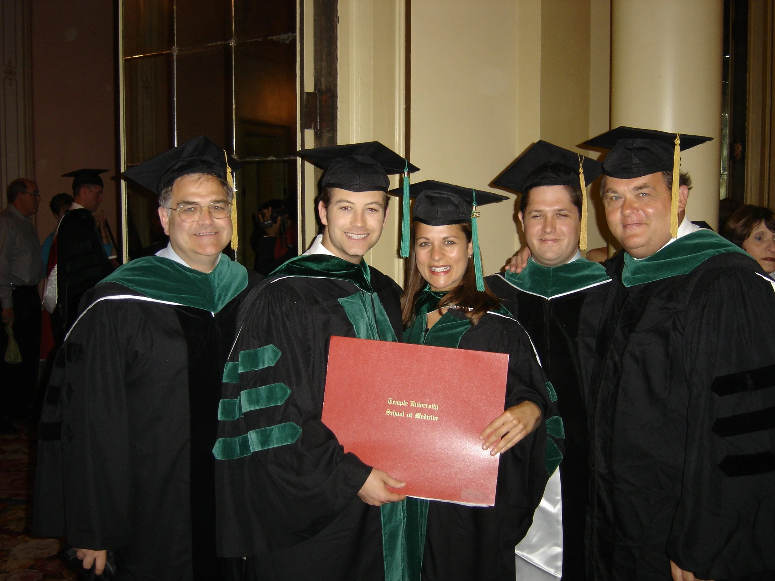 Group of people in graduation caps and gowns standing indoors, celebrating graduation ceremony, woman holding a diploma from Temple University.