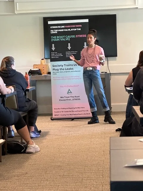 A woman in pink shirt and jeans giving a presentation in a classroom with students seated at desks, a large screen behind her, and a pink and white poster about plumbing leaks.