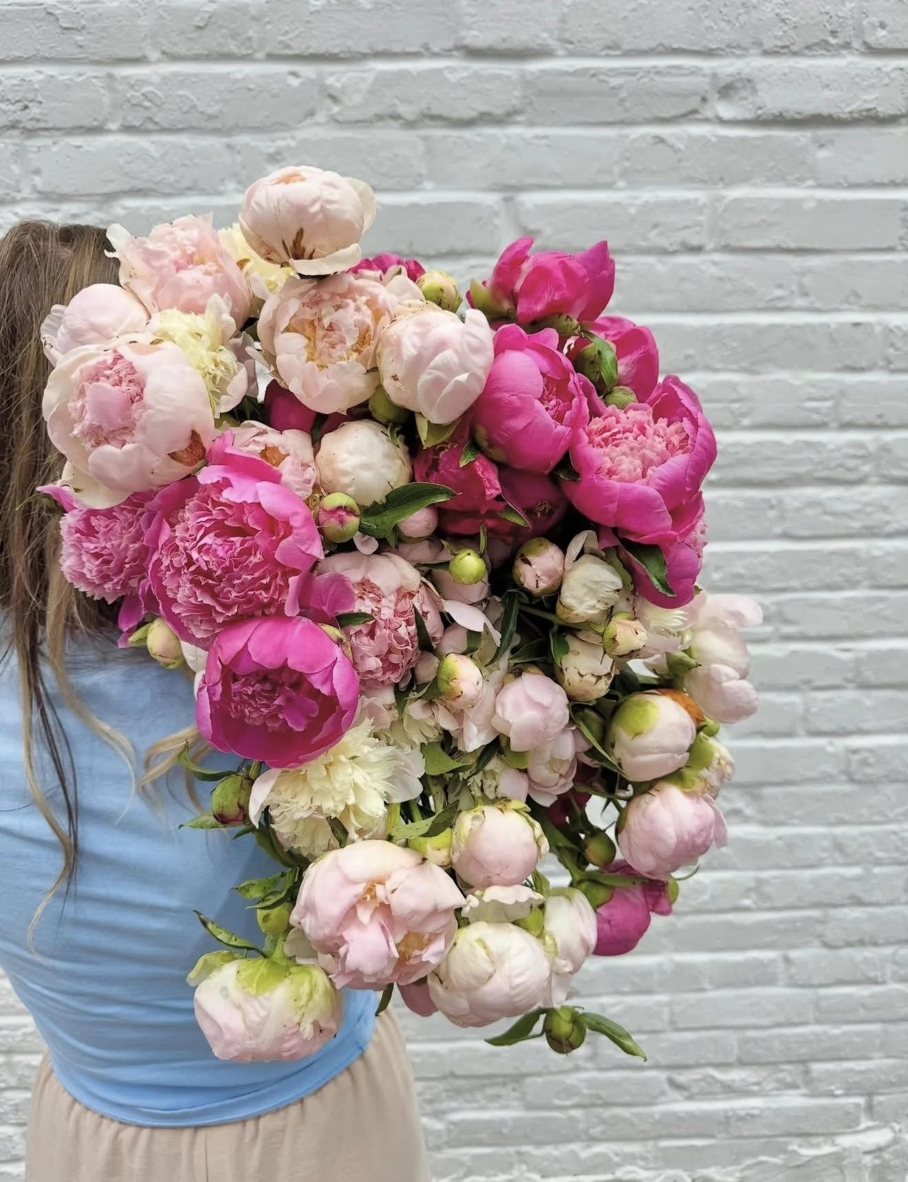 Person holding a lush bouquet of pink and blush peonies against a white brick wall.