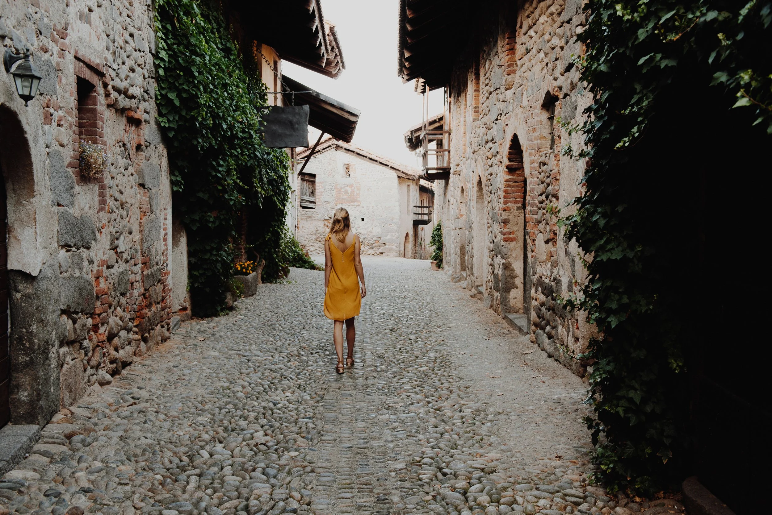 woman with blonde hair wearing a yellow-orange sundress walking down a cobblestone path road flanked by old walls covered in ivy in what looks like an old Italian village