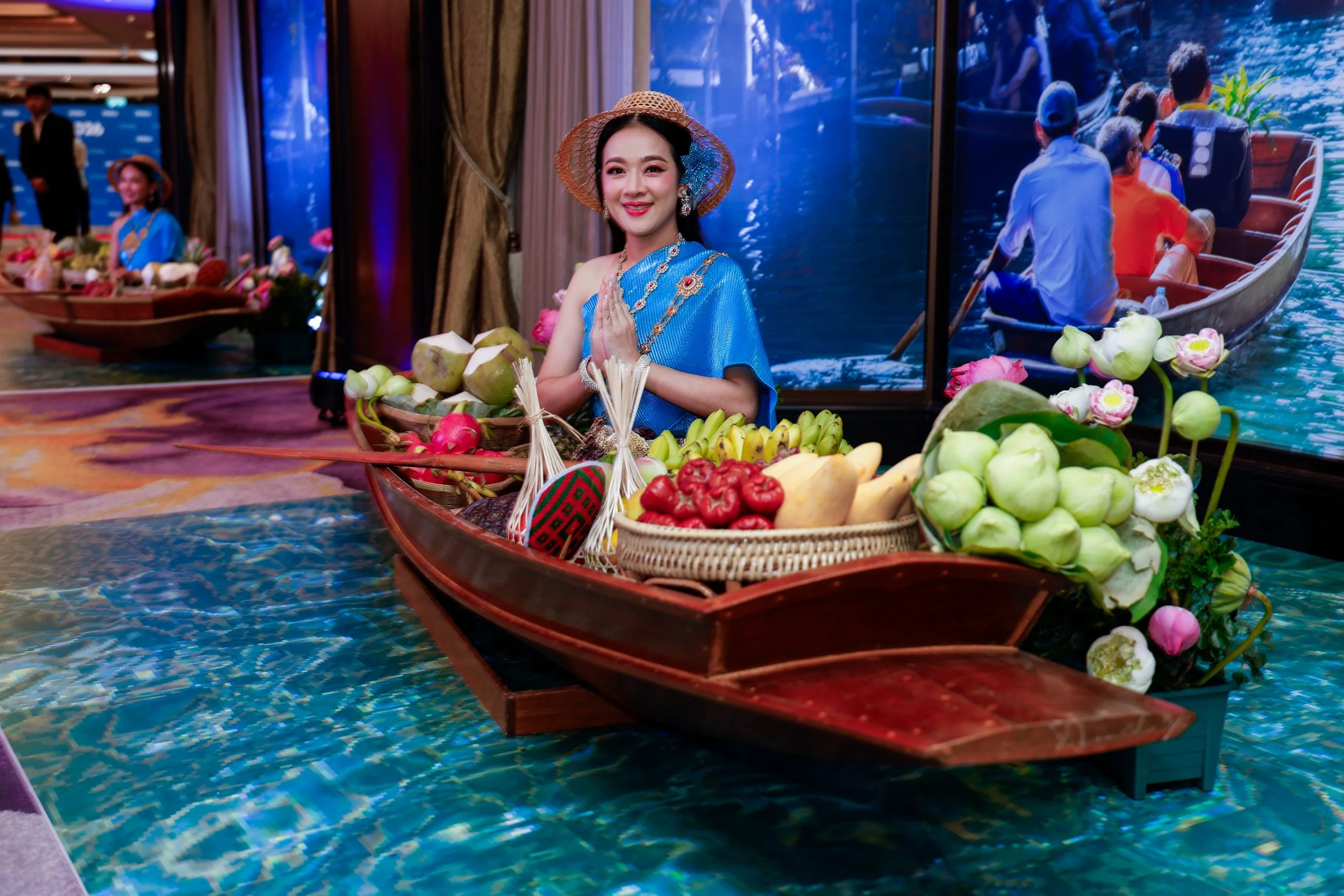 A woman in traditional Asian attire and jewelry standing behind a boat filled with fresh fruits, with a backdrop of water and boat images on the wall