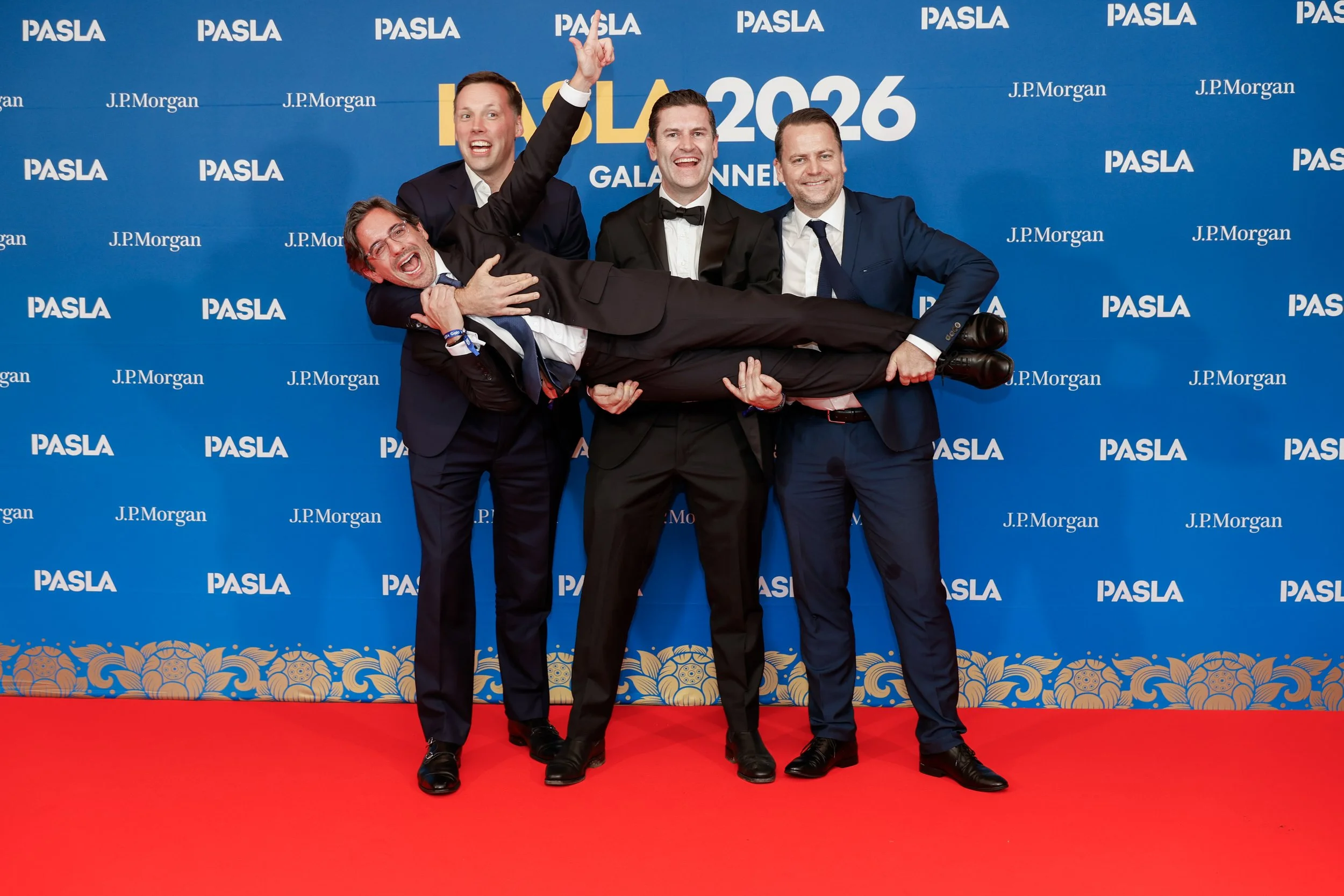 Four men dressed in formal tuxedos posing on a red carpet at the NASLA 2026 Gala Dinner with a blue backdrop displaying the event logos sponsored by J.P. Morgan, celebrating their achievement and having fun.