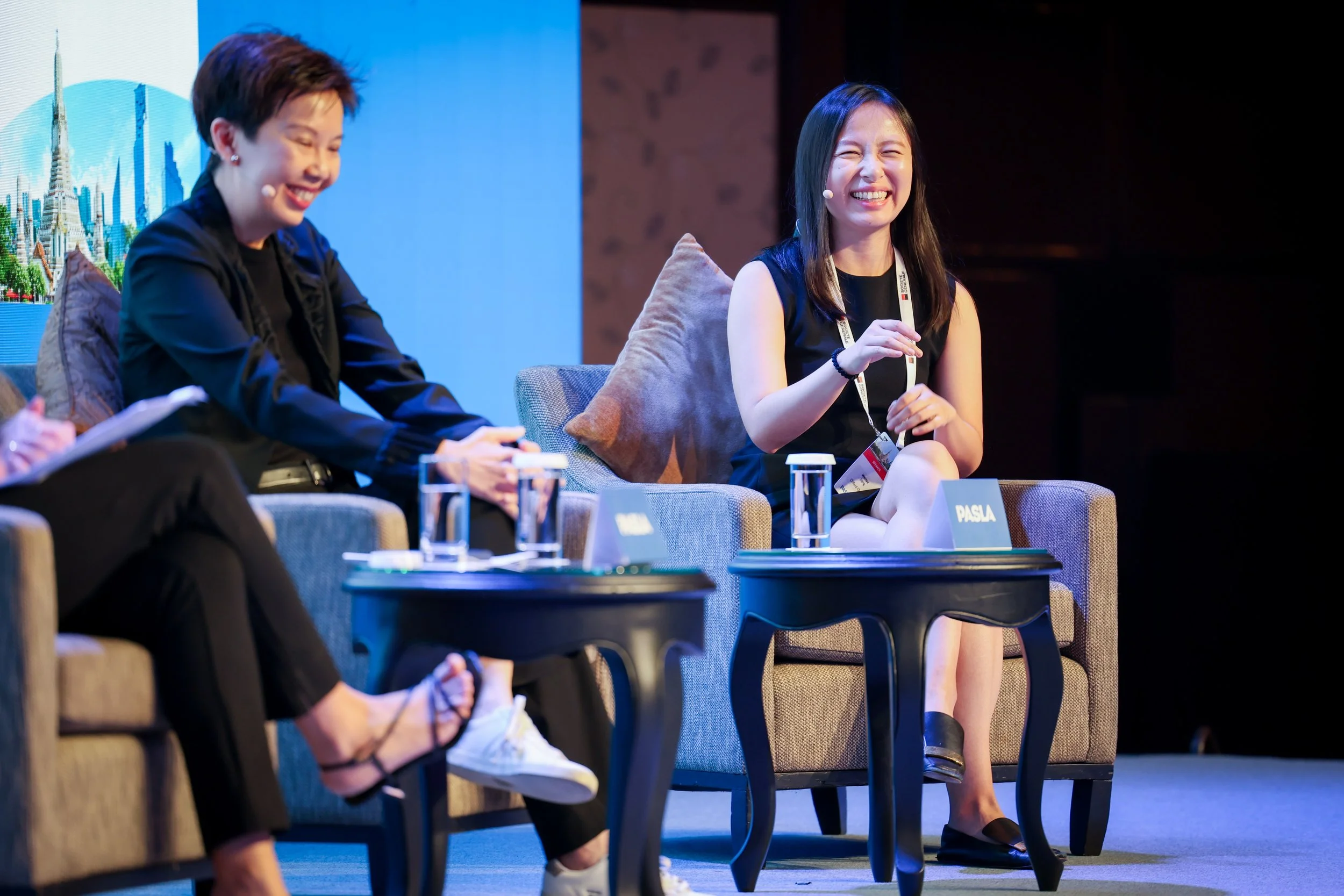 Two women sitting on stage, smiling and wearing microphones. One woman has short hair and the other has long hair. They are inside a conference room or event space with a large screen behind them.